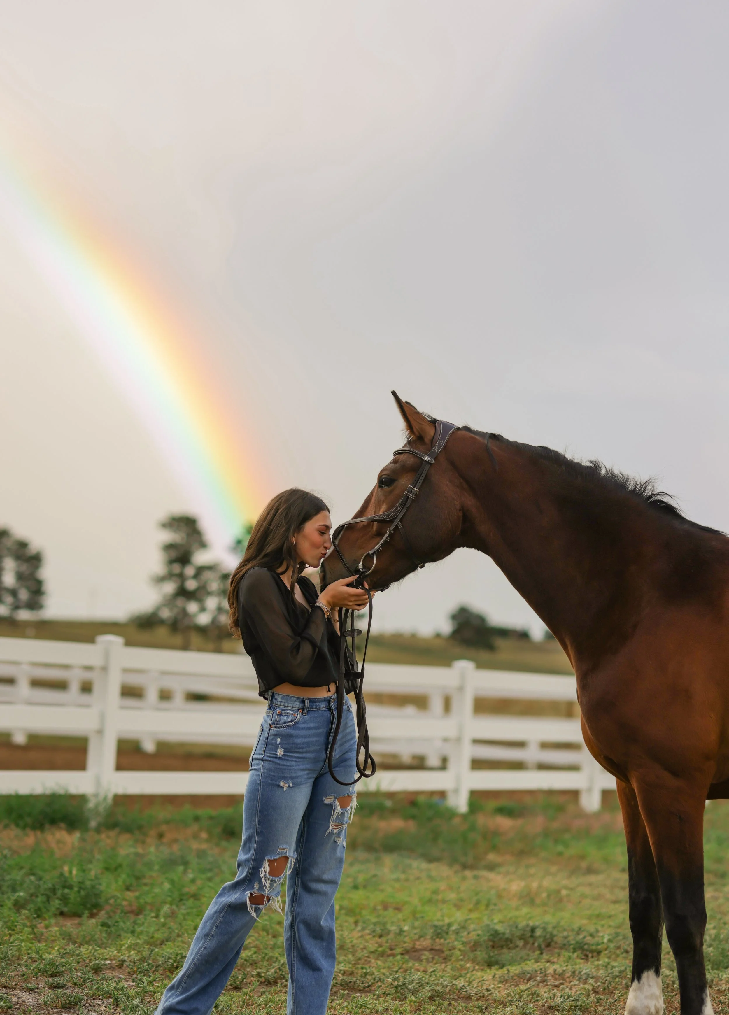 A young woman with long hair, wearing a black jacket and ripped jeans, holding a horse's bridle and kissing the horse on the nose in a grassy outdoor area with a white fence and trees in the background. A rainbow is visible in the cloudy sky.