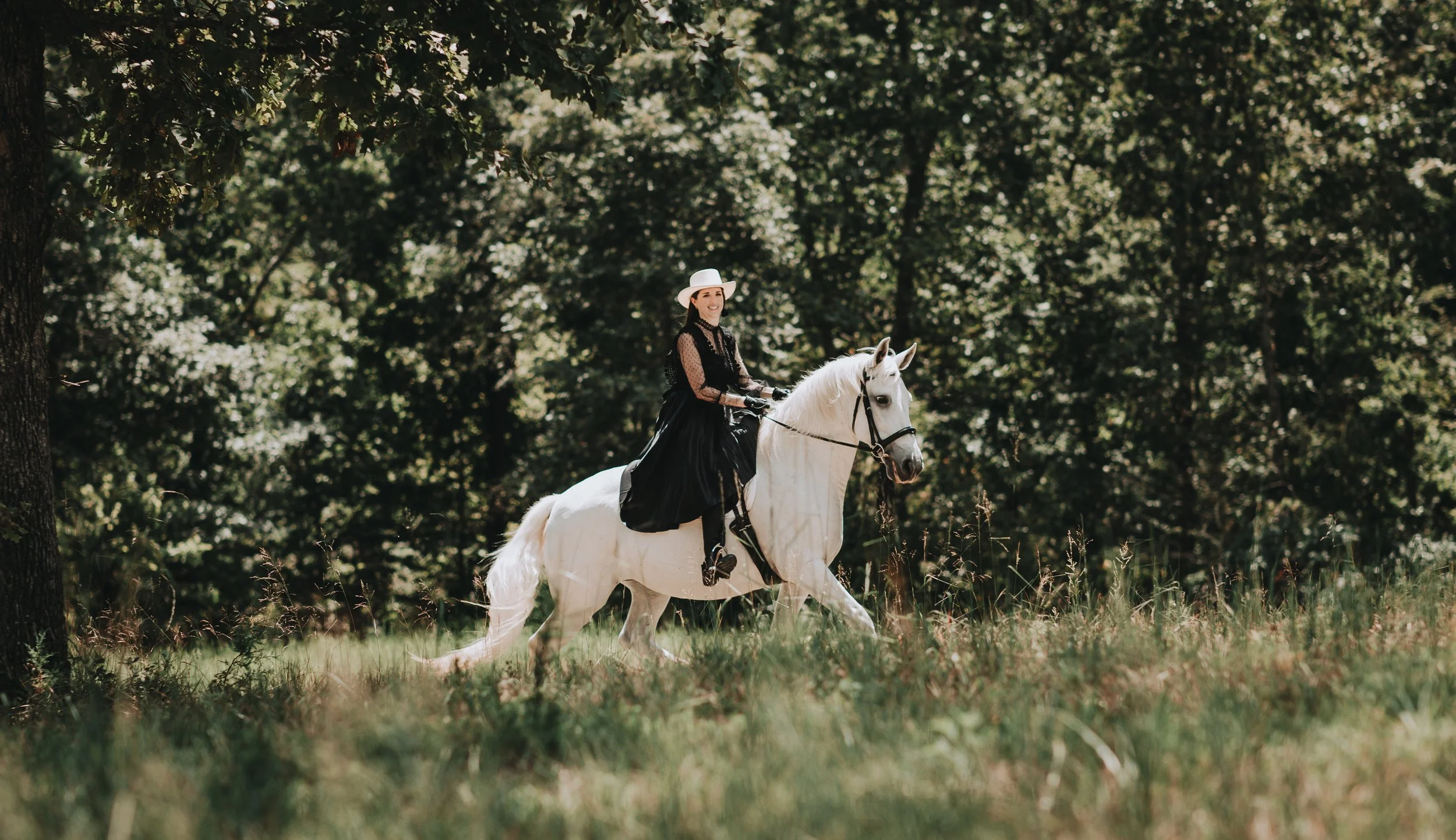 A woman wearing a black dress and a white hat riding a white horse through a green forested area.