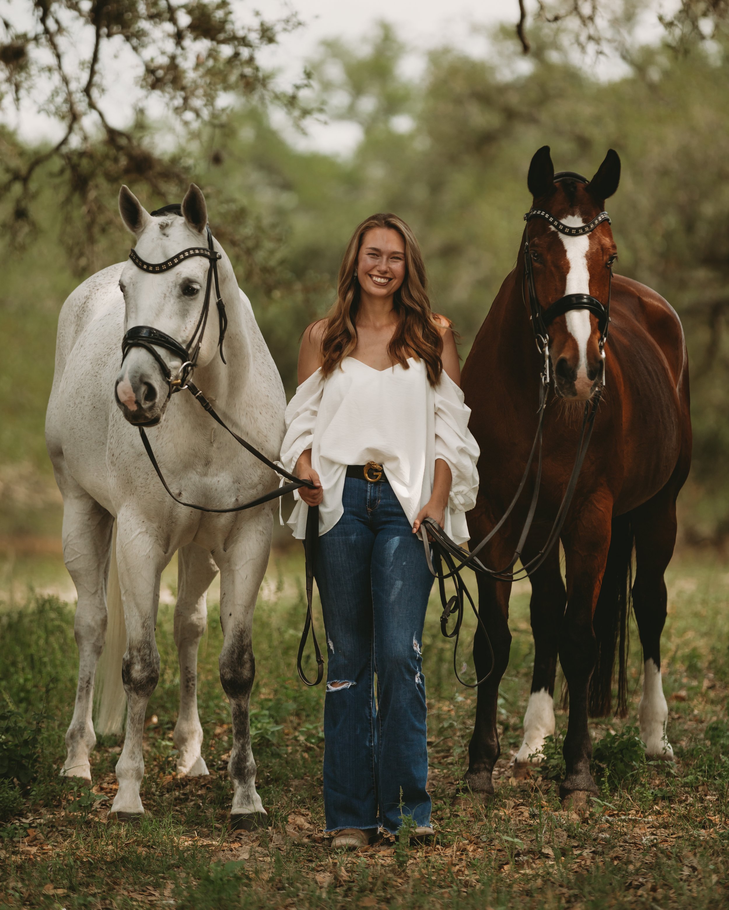 A woman standing outdoors between two horses, one white and one brown, with trees in the background, smiling.