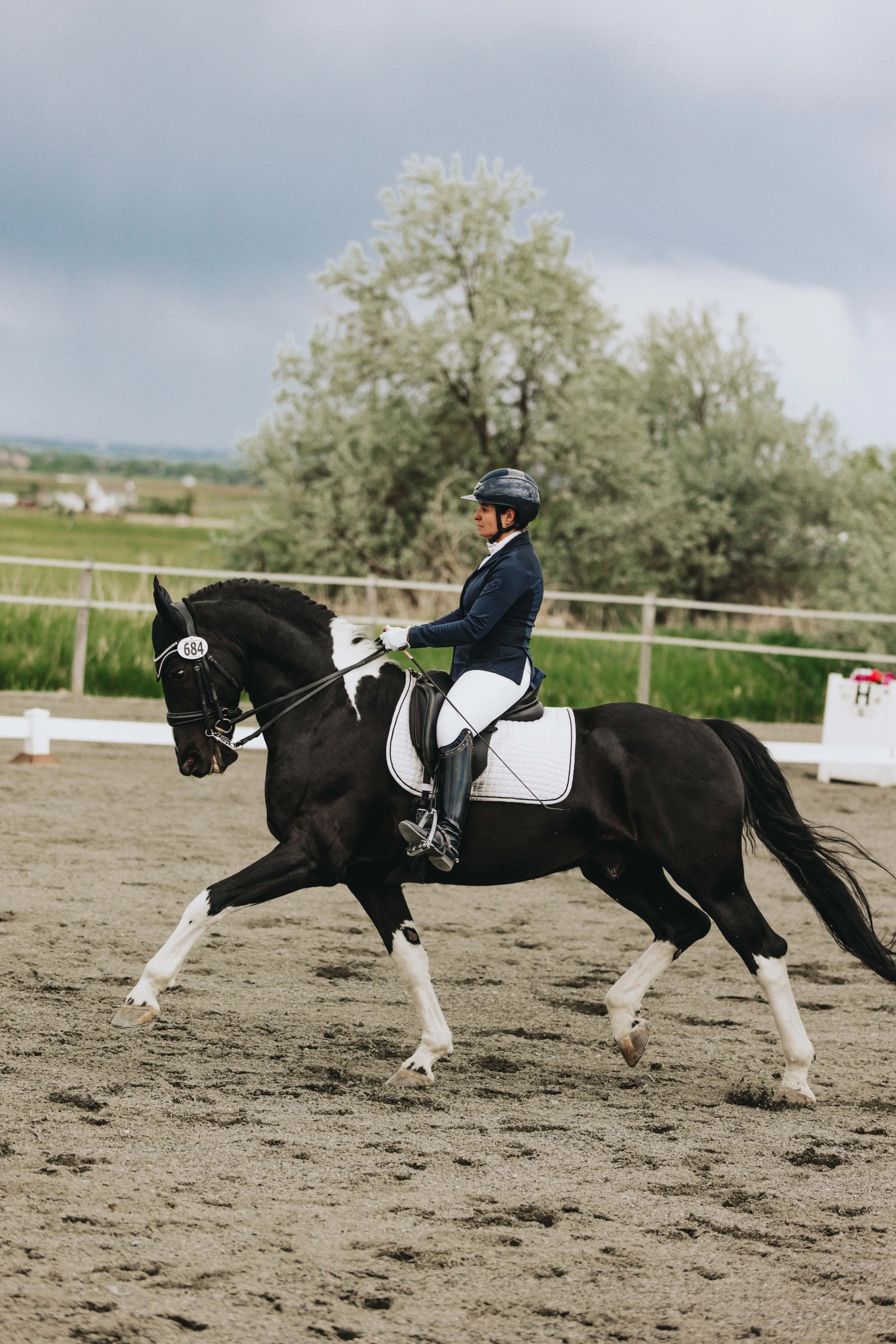 A female equestrian riding a black and white pinto horse during a dressage competition outdoors on a cloudy day.