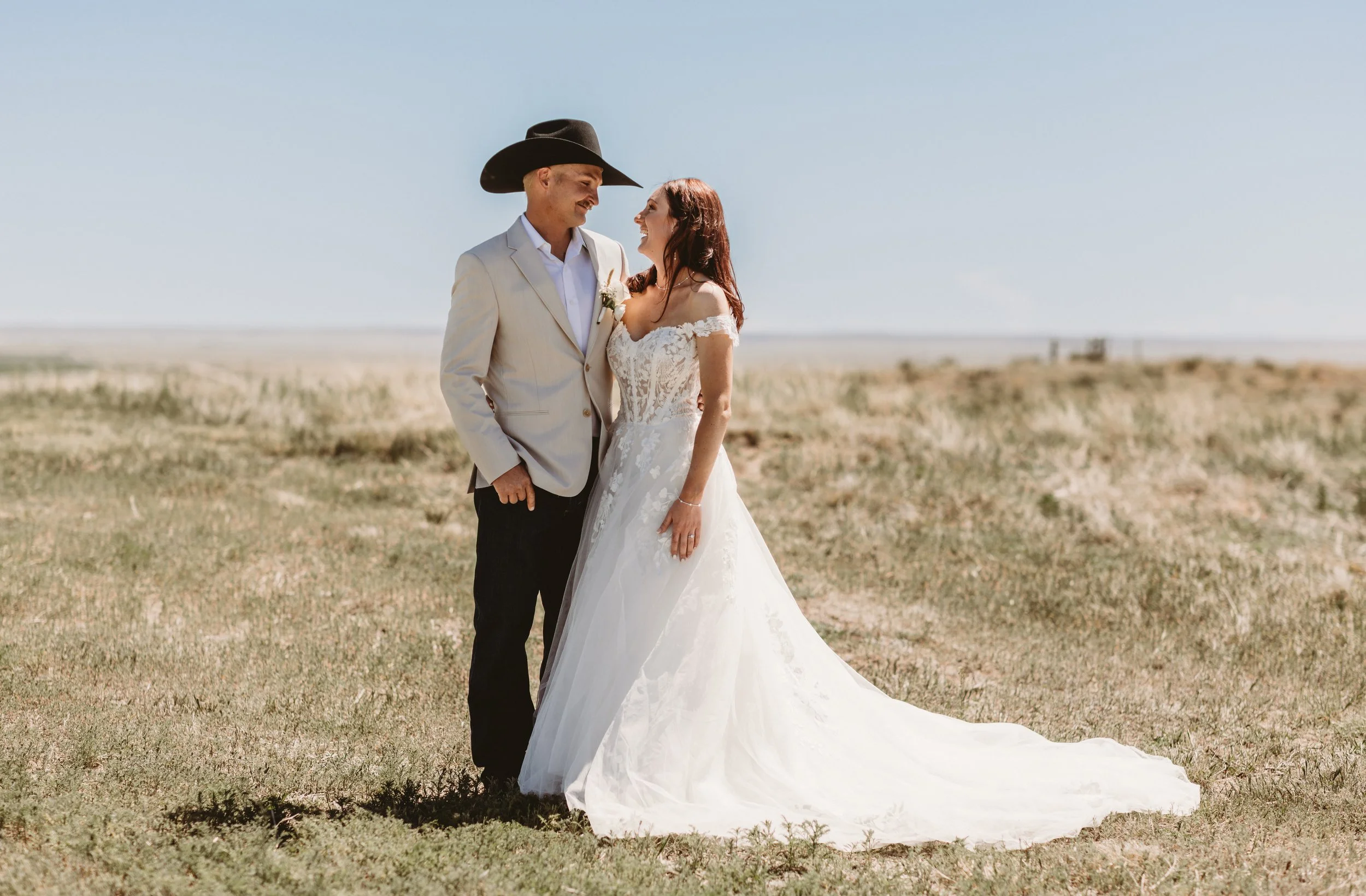 A couple in wedding attire standing closely on a desert-like landscape with a clear blue sky, smiling at each other.