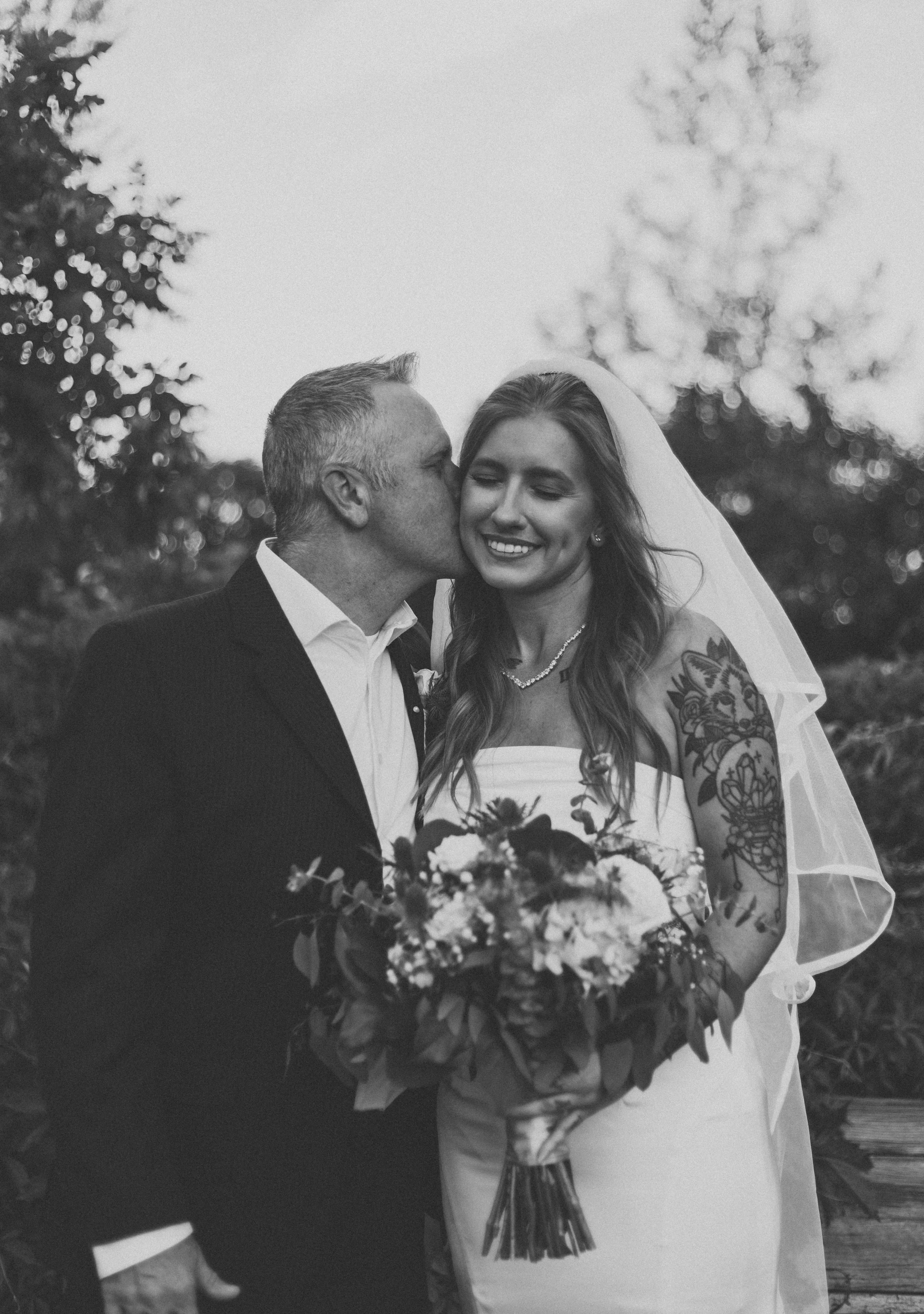 Black and white photo of a bride and groom on their wedding day, with the groom kissing the bride's cheek. The bride is smiling, wearing a wedding dress and veil, holding a bouquet of flowers. The groom is in a suit. Out-of-focus trees are in the bac