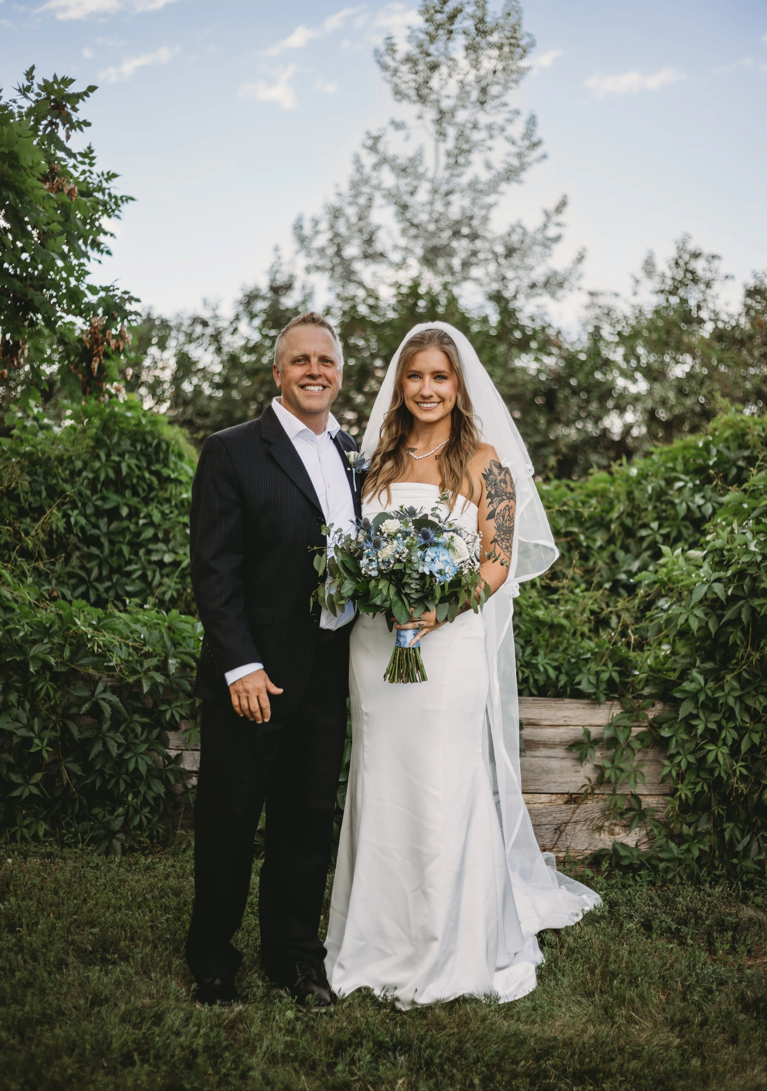 A man and woman in wedding attire posing outdoors with greenery and a cloudy sky in the background.