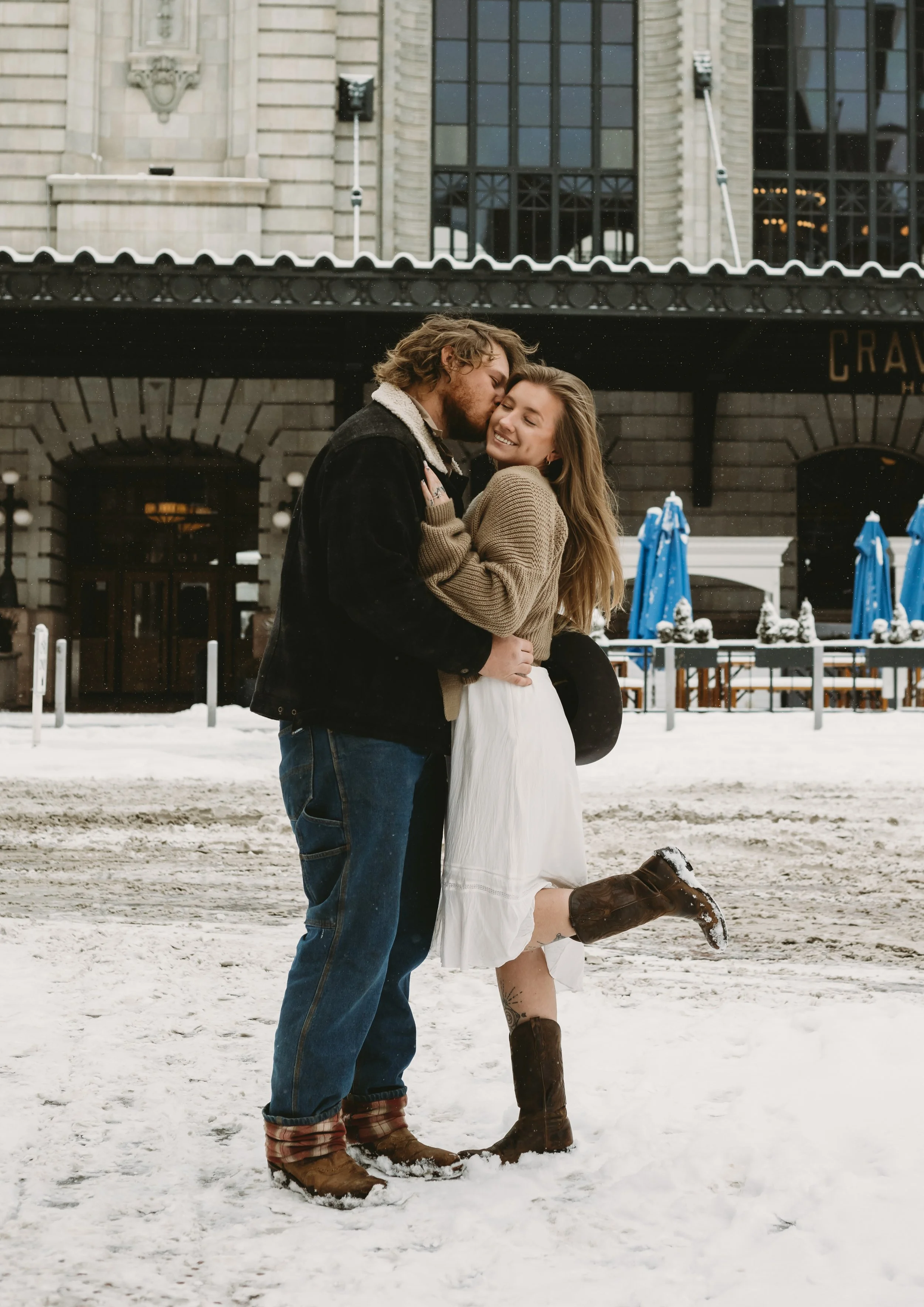 A smiling woman and man embrace in the snow in front of a historic building with large windows and ornate architectural details. The woman is kneeling on one knee, wearing a white dress and brown boots, while the man is standing, wearing jeans, a jac