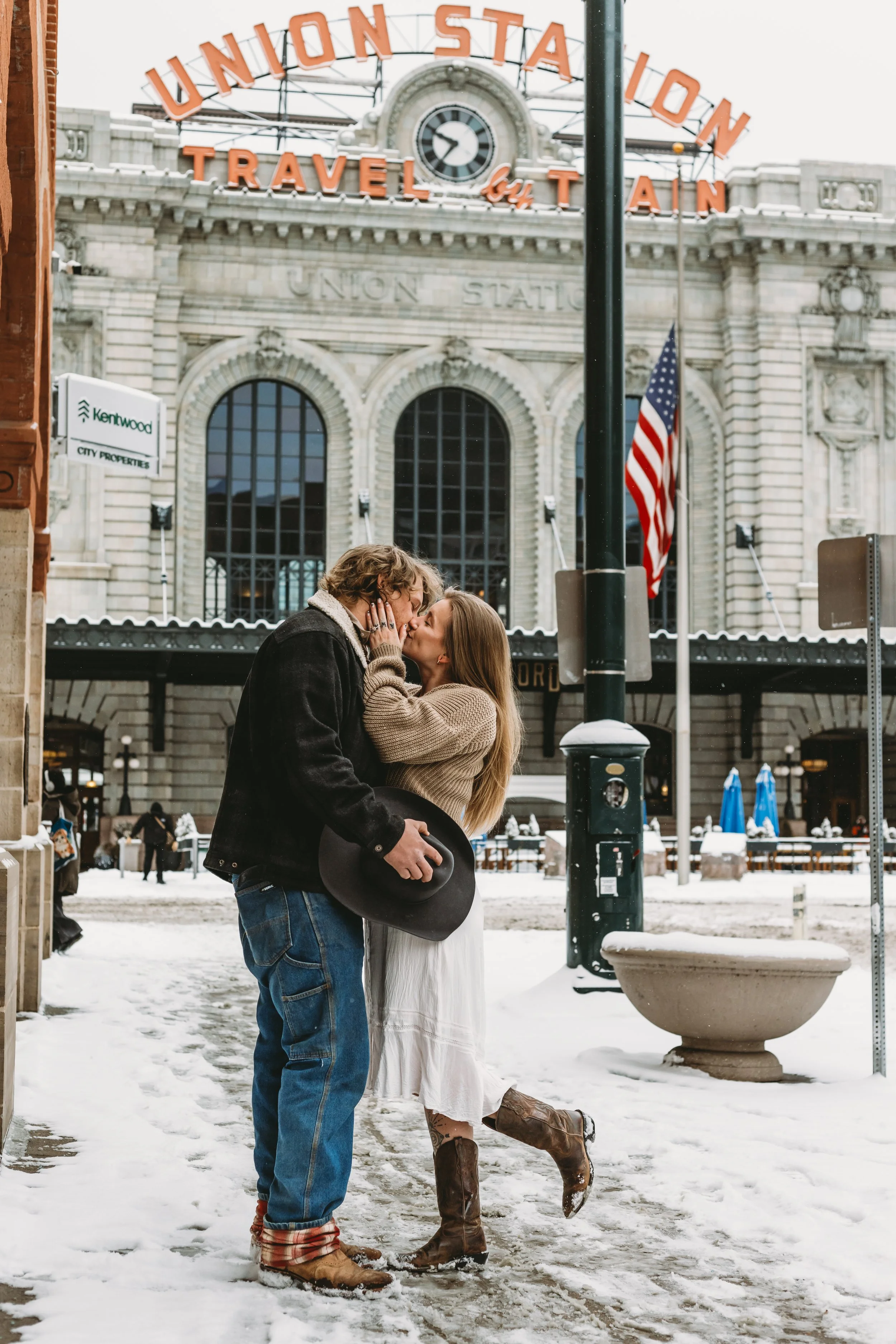 A couple shares a romantic kiss in front of Union Station, with snow on the ground and people strolling in the background.