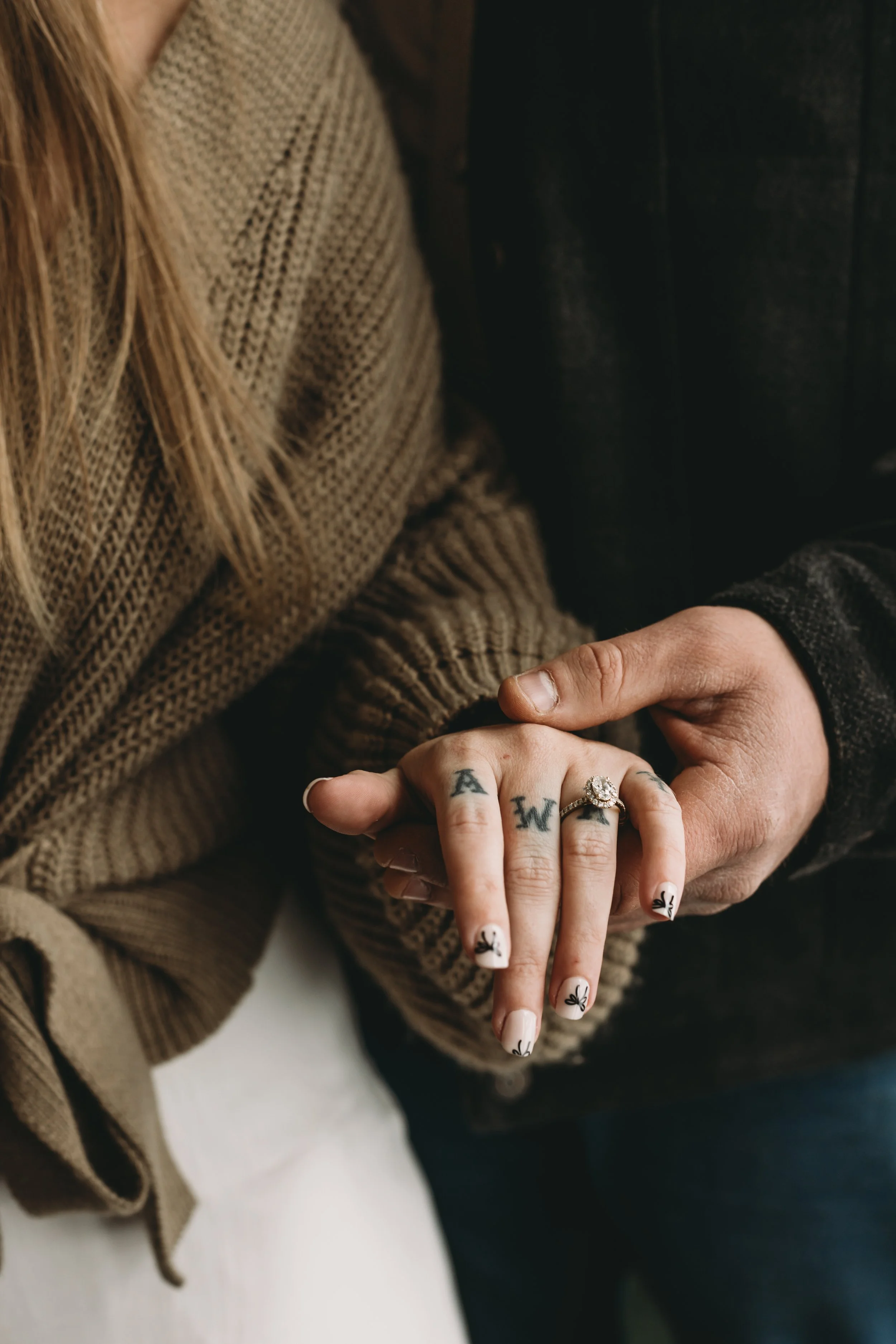 Close-up of a woman with long blonde hair, wearing a brown knit sweater, holding hands with a man. The woman's nails have tattoos of bees and butterflies, and she wears rings on her fingers.