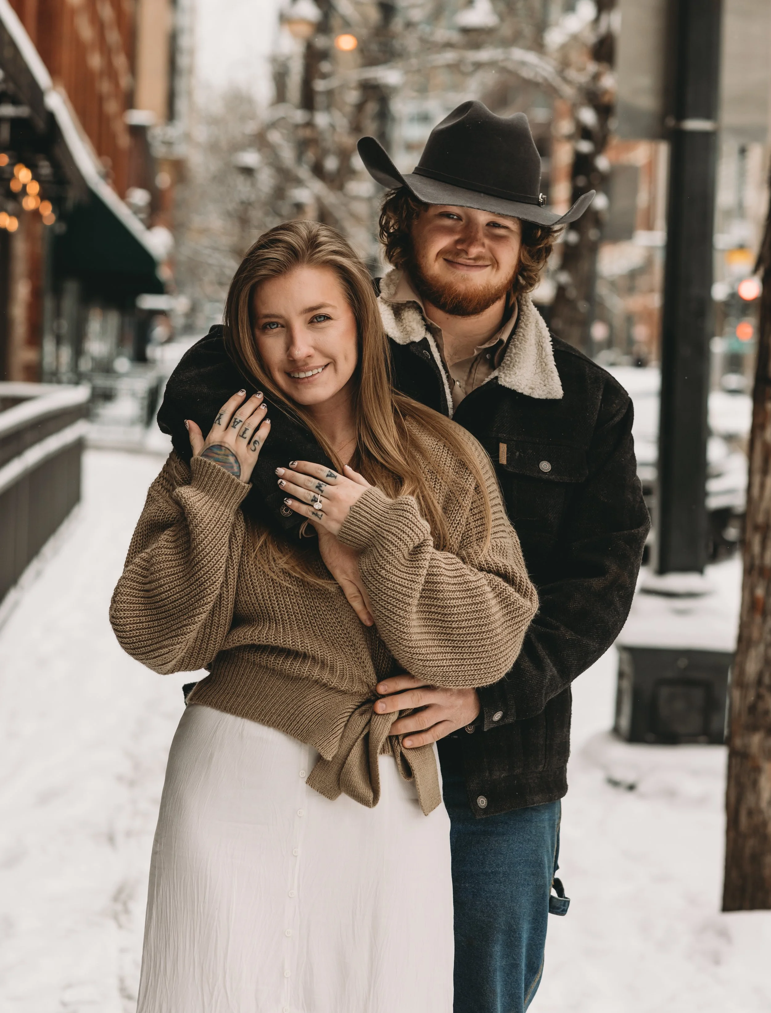 A young couple is standing close together outside on a snowy street, smiling and embracing each other. The woman has long light brown hair, wearing a brown sweater, white skirt, and has tattoos on her hands. The man has curly hair, a beard, wearing a