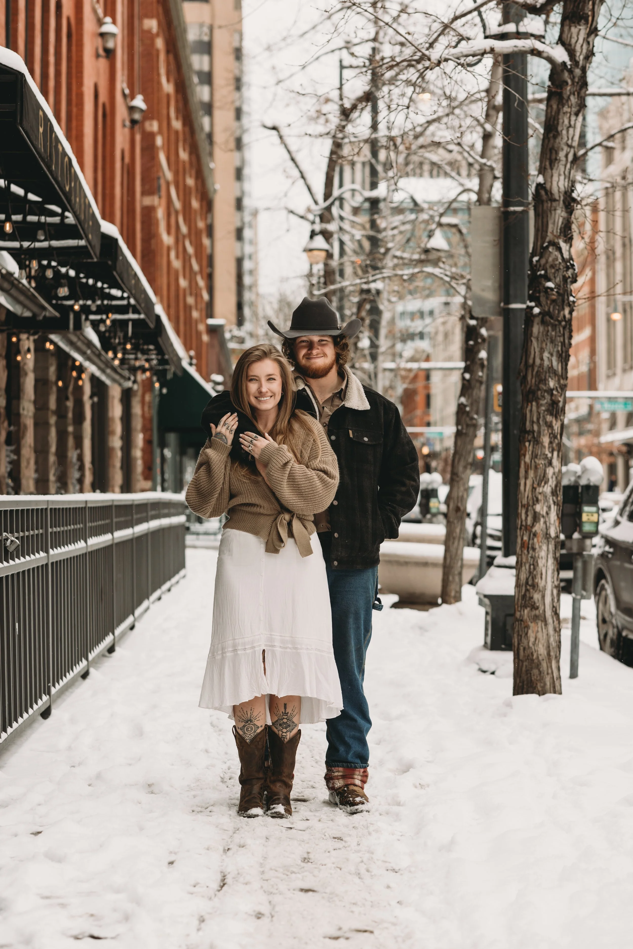 A smiling young woman and a man with a cowboy hat walk arm-in-arm on a snowy city sidewalk, with leafless trees and brick buildings in the background.