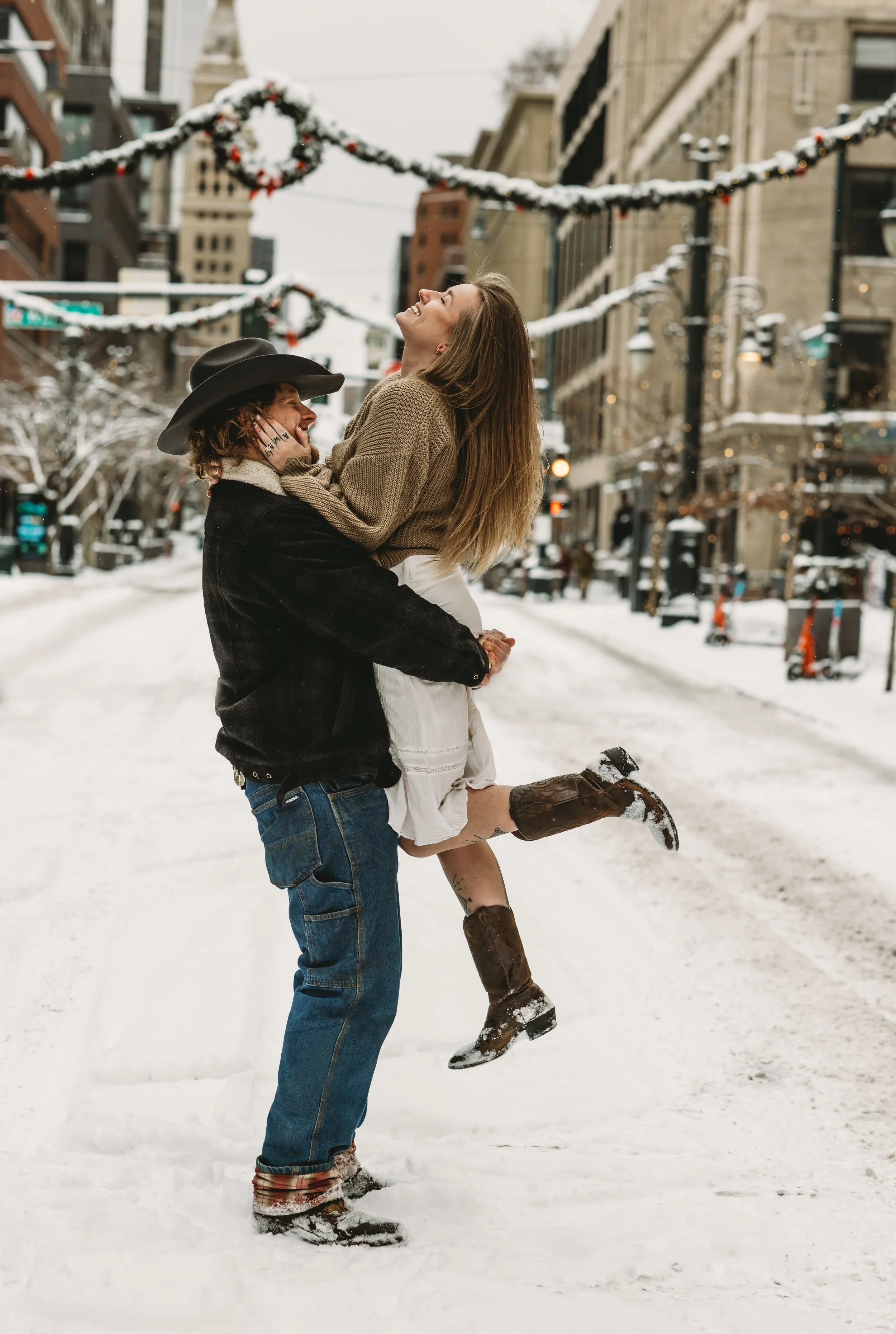A man wearing jeans, a black jacket, and a cowboy hat lifting a woman in a beige sweater and white skirt in a snowy city street decorated with holiday garlands.