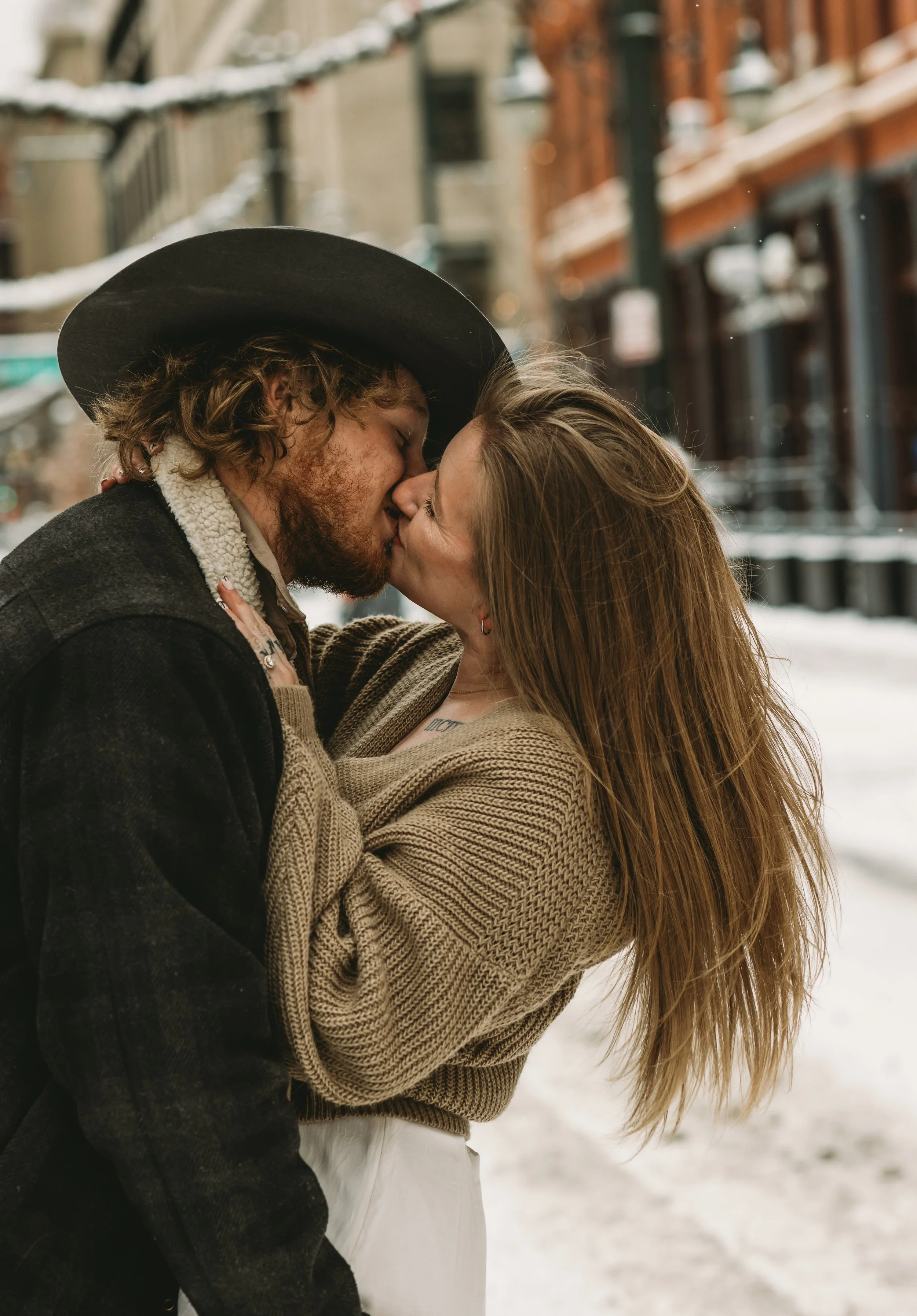 A couple is kissing outdoors in a snowy urban setting, with the man wearing a wide-brimmed hat and the woman with long red hair, both dressed warmly.