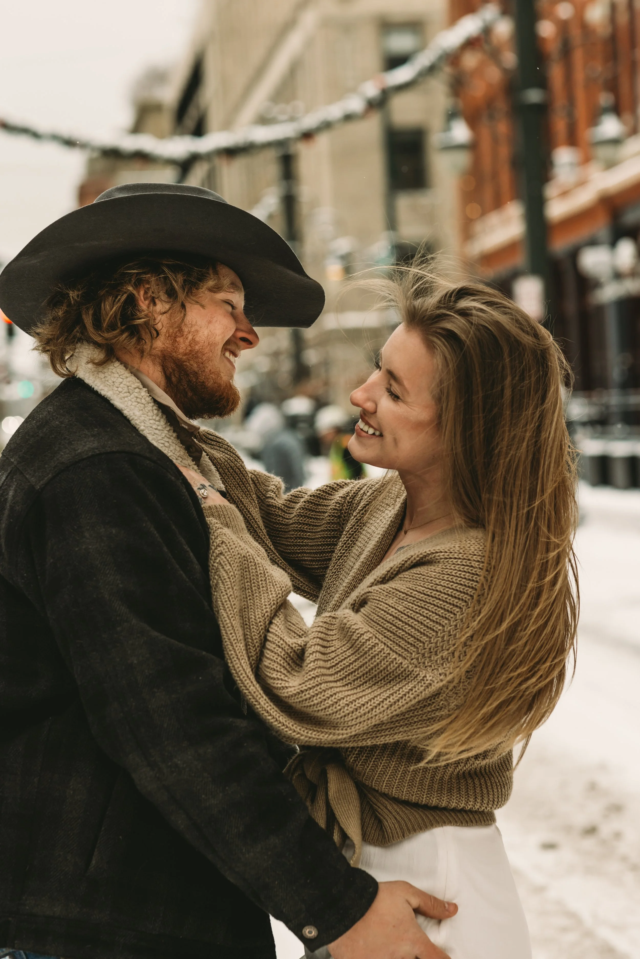 A man and woman are smiling and looking at each other in an outdoor urban setting in winter. The man is wearing a large wide-brimmed hat and a dark coat, while the woman has long blonde hair and is wearing a beige sweater.