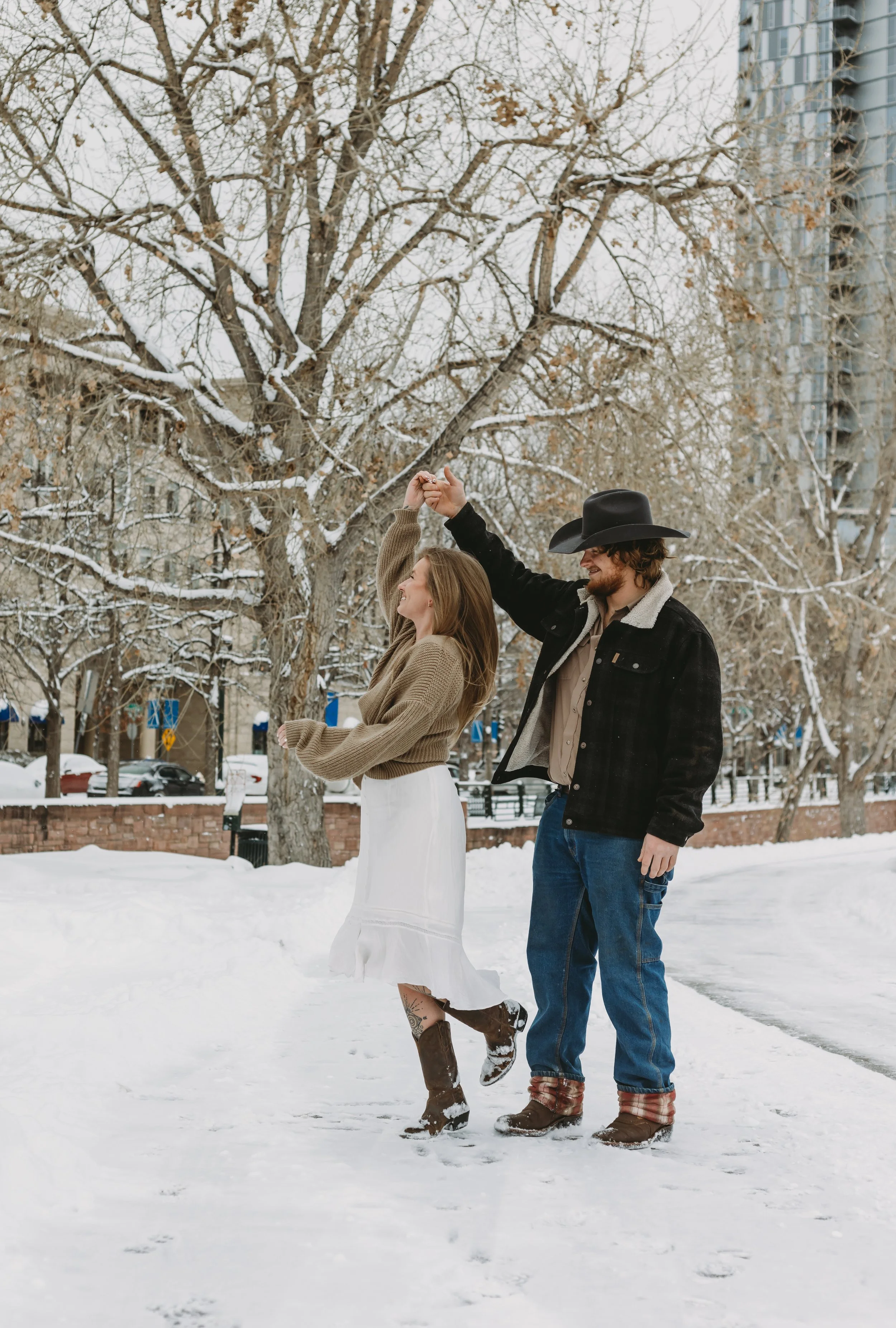 A couple dancing in the snow outdoors, holding hands, with snow-covered trees and buildings in the background.