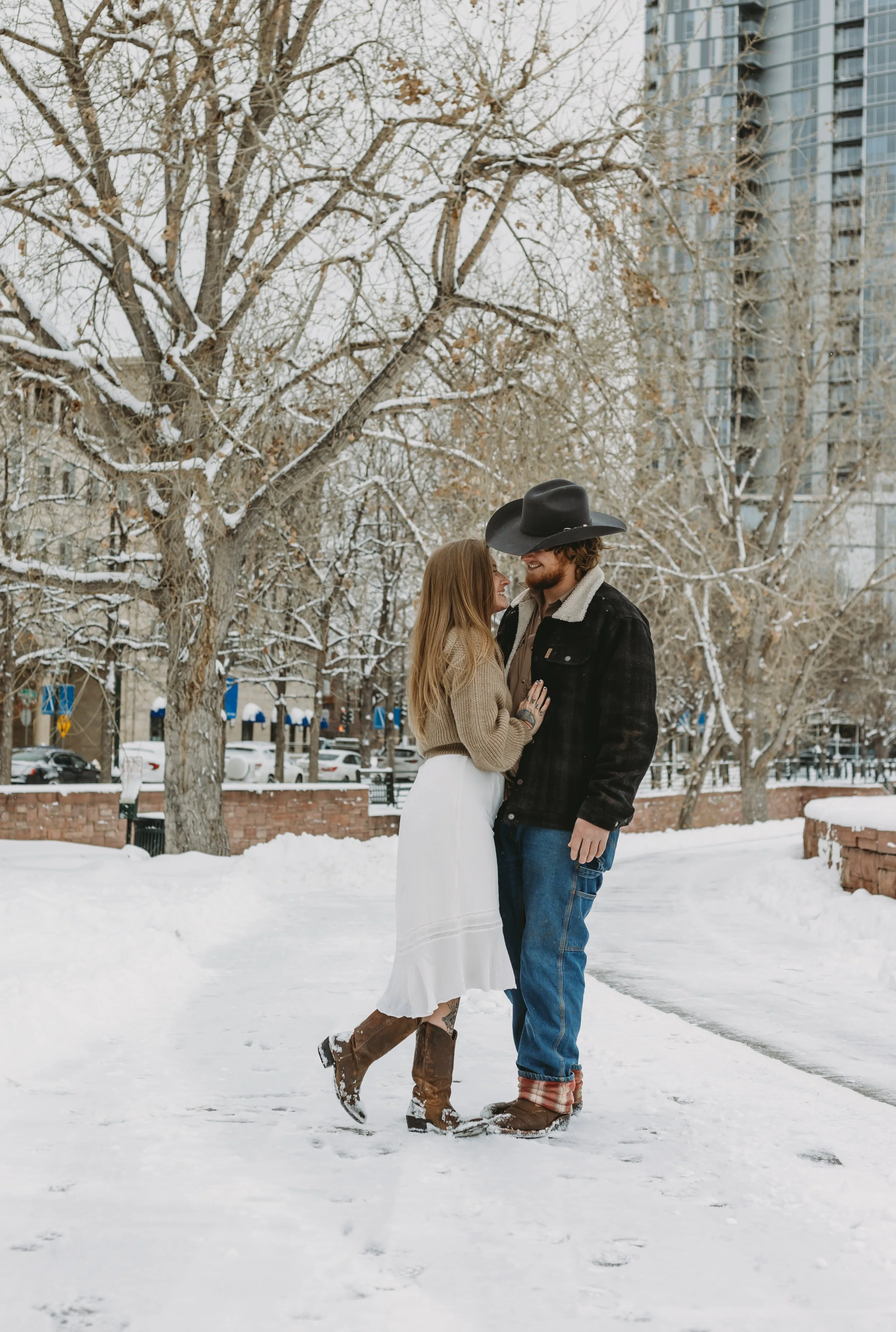A couple stands close and smiling in a snowy park with leafless trees and tall buildings in the background. The woman is wearing a beige sweater, white skirt, and brown boots, while the man is dressed in a cowboy hat, dark jacket, and jeans.