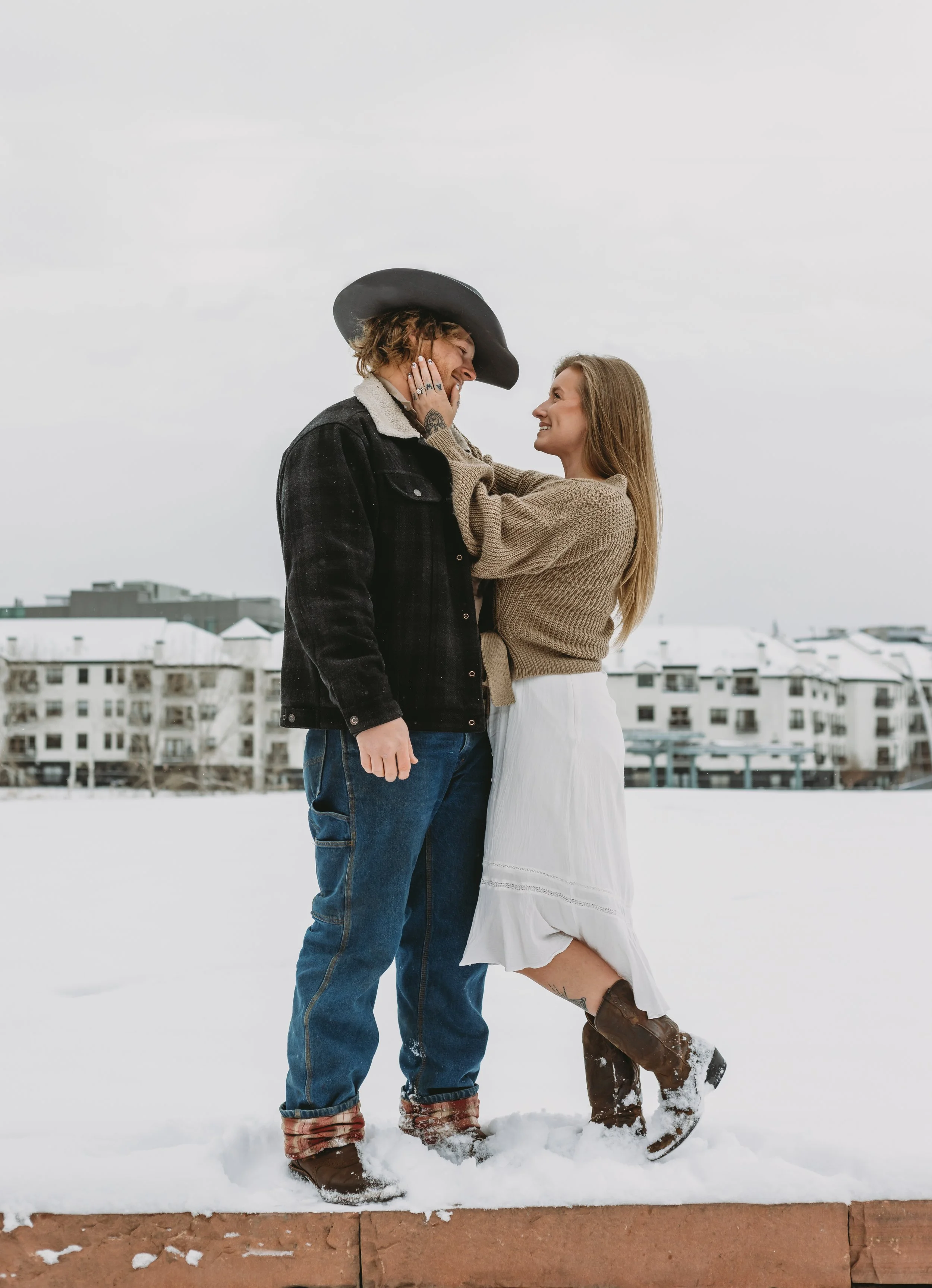 A man and a woman stand close together in a snowy outdoor setting, smiling and looking at each other, with the man touching the woman's face. The man is wearing a black jacket, jeans, and a wide-brimmed black hat, while the woman is wearing a tan swe
