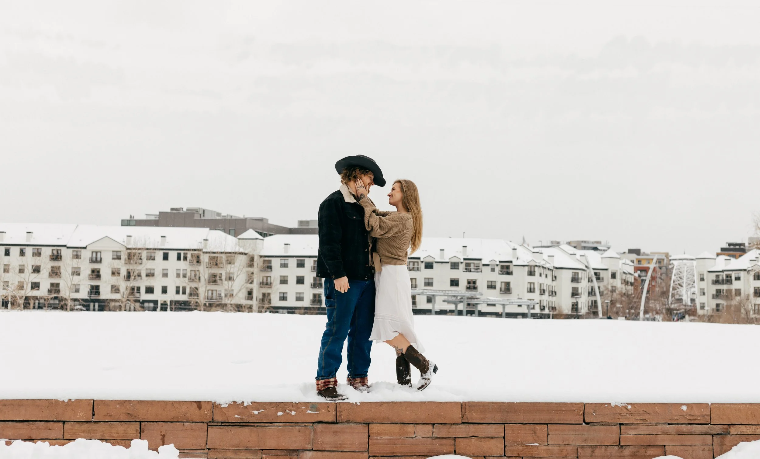 A couple standing close together in a snowy urban area, smiling at each other, with modern apartment buildings in the background.
