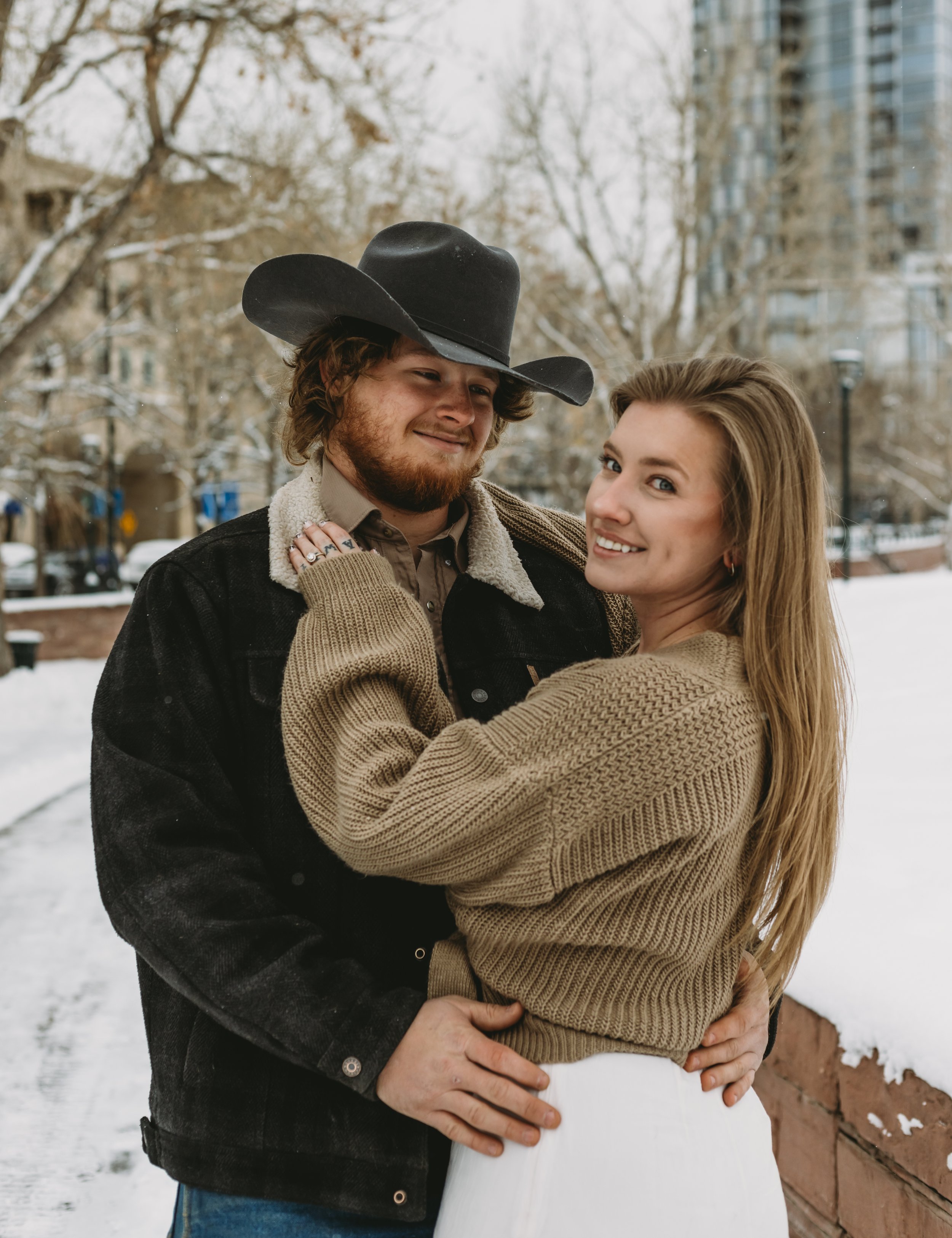 A smiling young woman embraces a man wearing a large black cowboy hat outdoors in a winter city park with snow on the ground, trees, and buildings in the background.