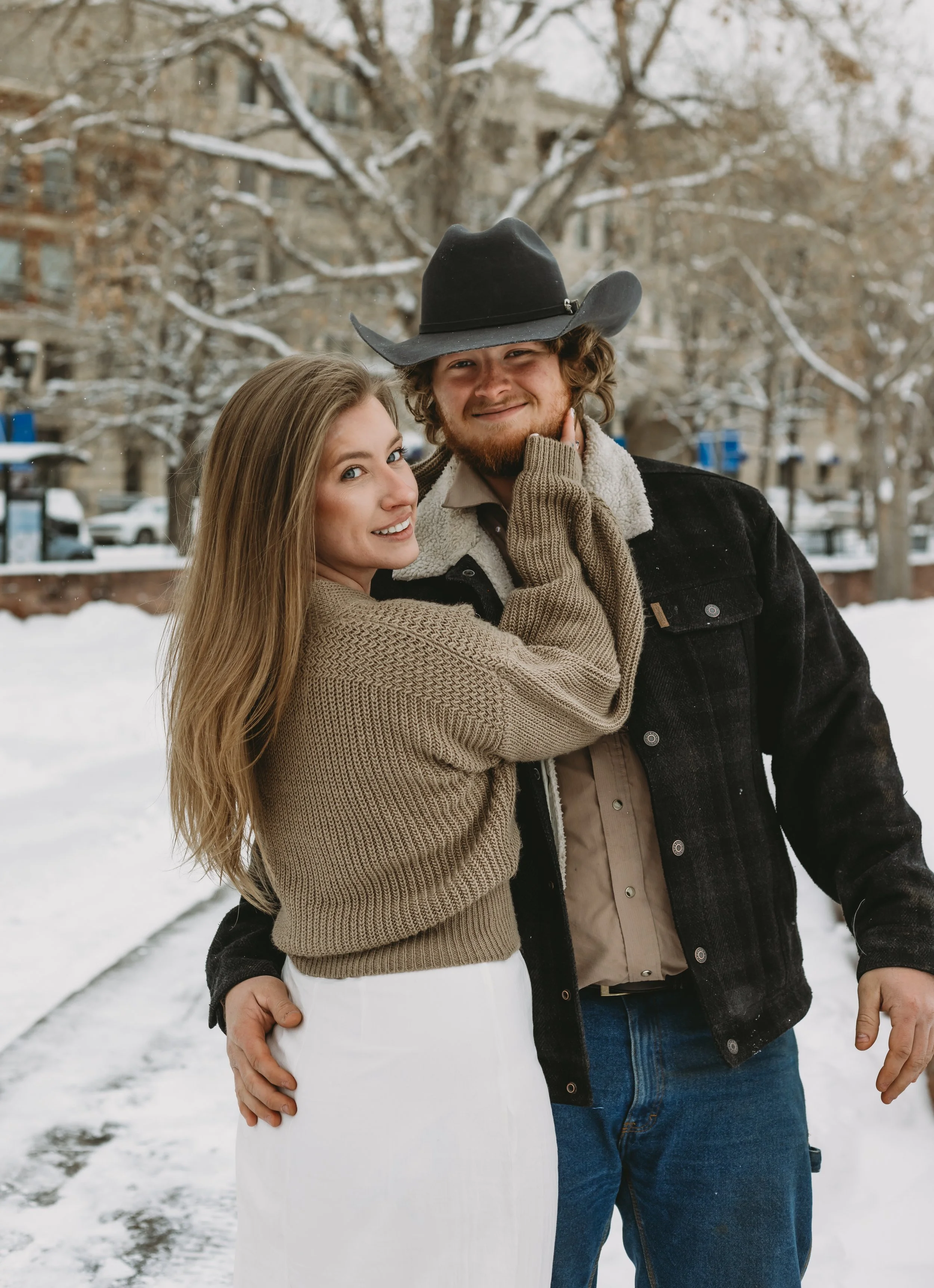 A couple is posing outdoors in a snowy park during winter. The woman has long blonde hair and is wearing a beige sweater with white pants. The man has curly hair, a beard, and is wearing a black cowboy hat, a black jacket with a shearling collar, and