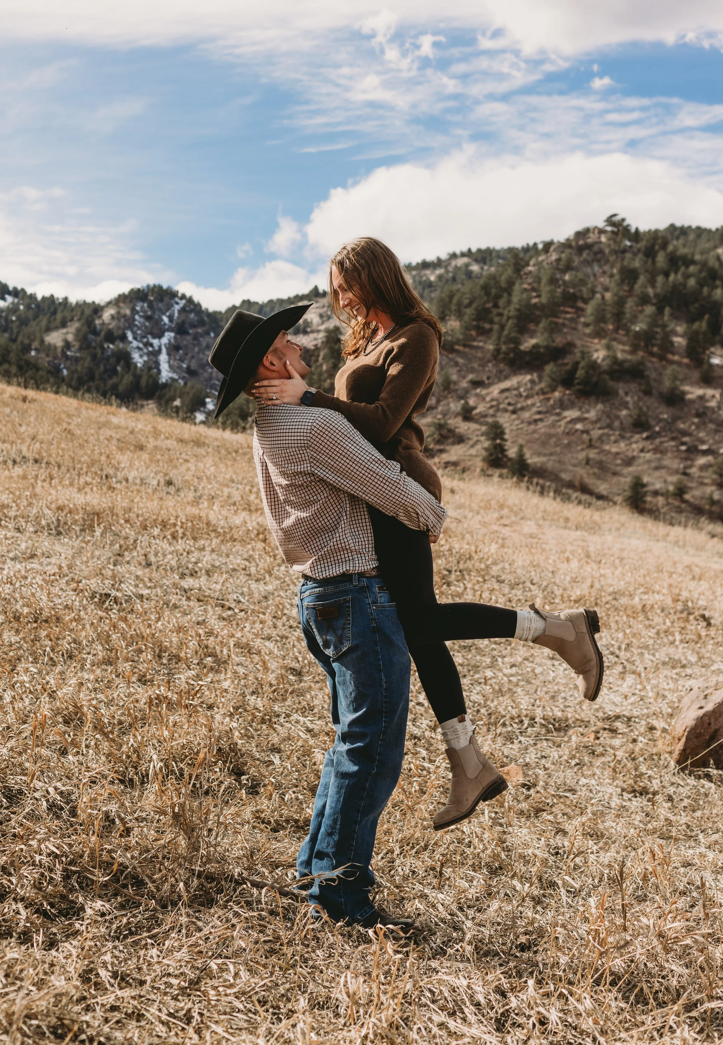 A couple in a field with mountains in the background: man lifting woman, both smiling and looking at each other.