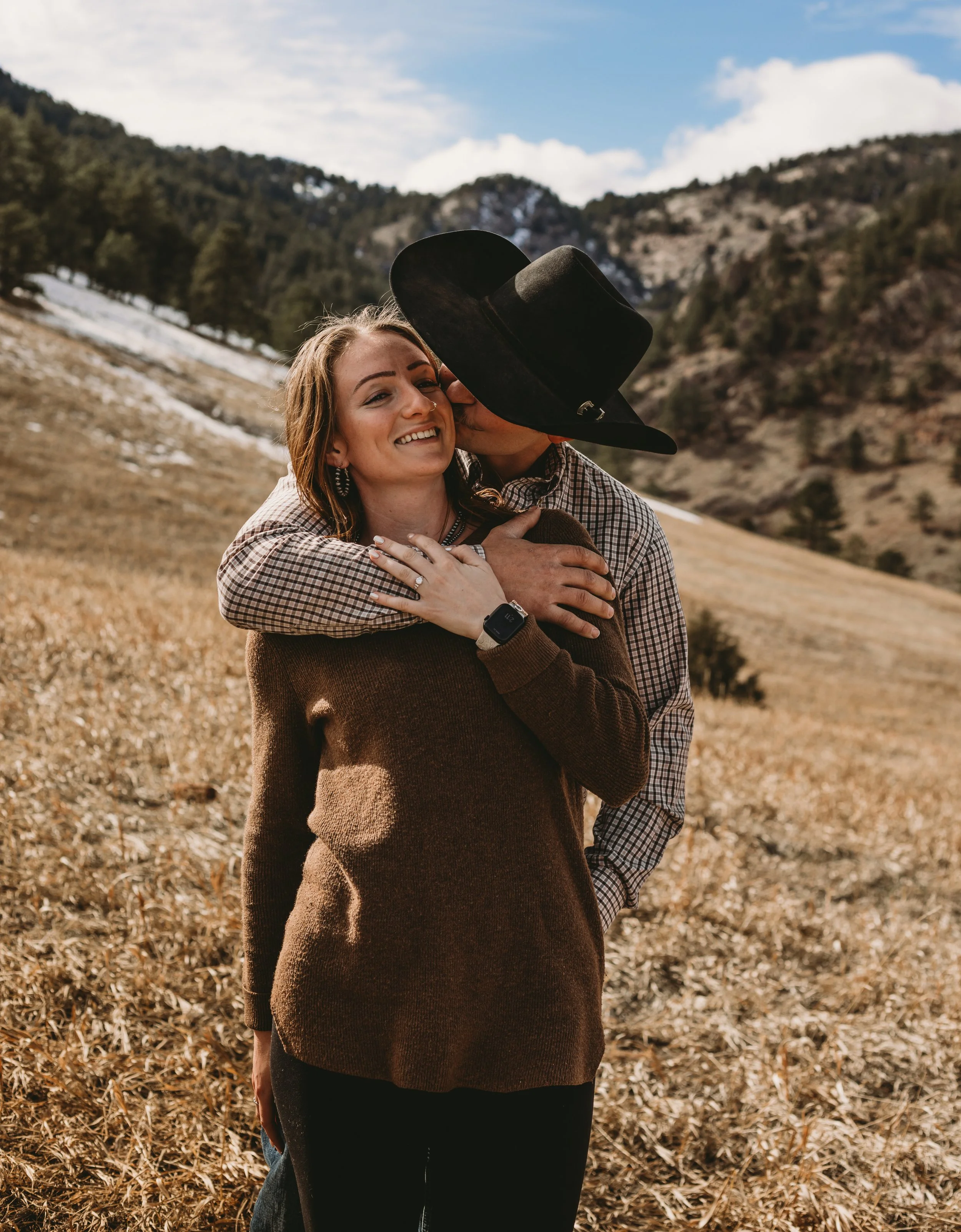 A man giving a woman a kiss on the cheek in an outdoor mountain field during the daytime.