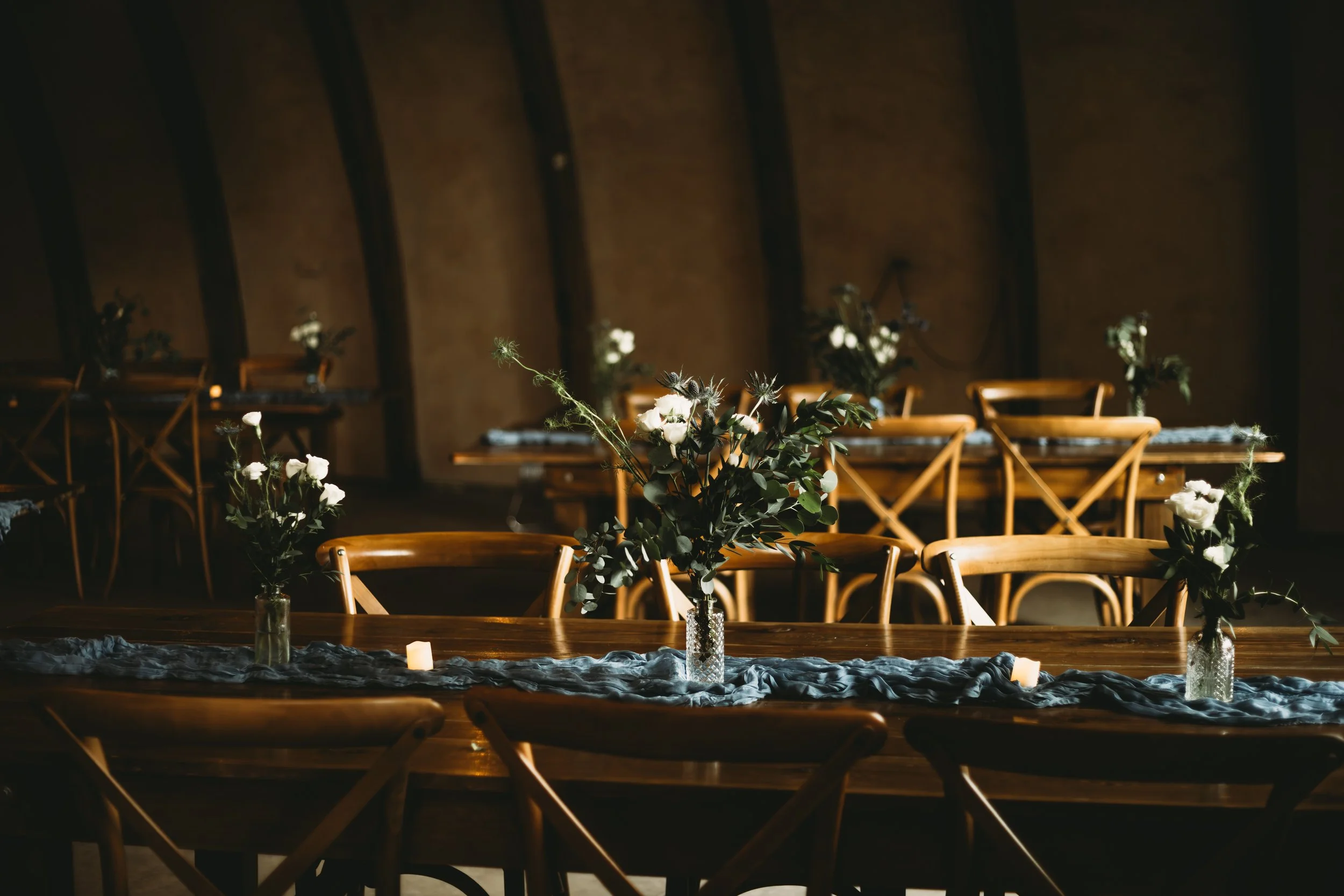 Wedding or event reception area with wooden chairs and multiple floral centerpieces in glass vases, set on a long table with decorative fabric and candles in a dimly lit room with wood-paneled walls.