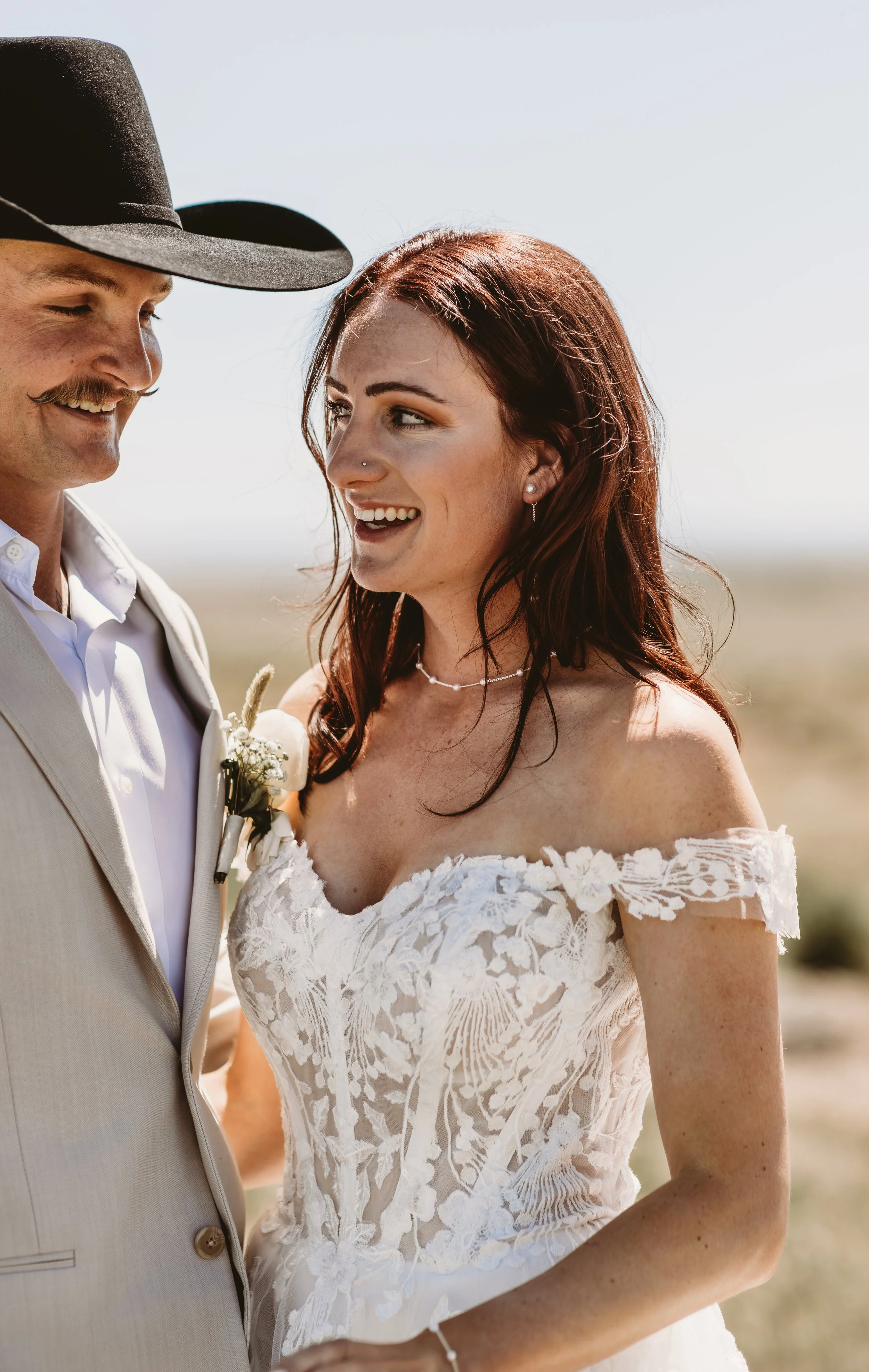 A bride and groom smile at each other outdoors on a sunny day. The bride has reddish-brown hair, wears a lace off-shoulder wedding dress, and has a nose piercing. The groom wears a light-colored suit and a black cowboy hat. They are close together, w