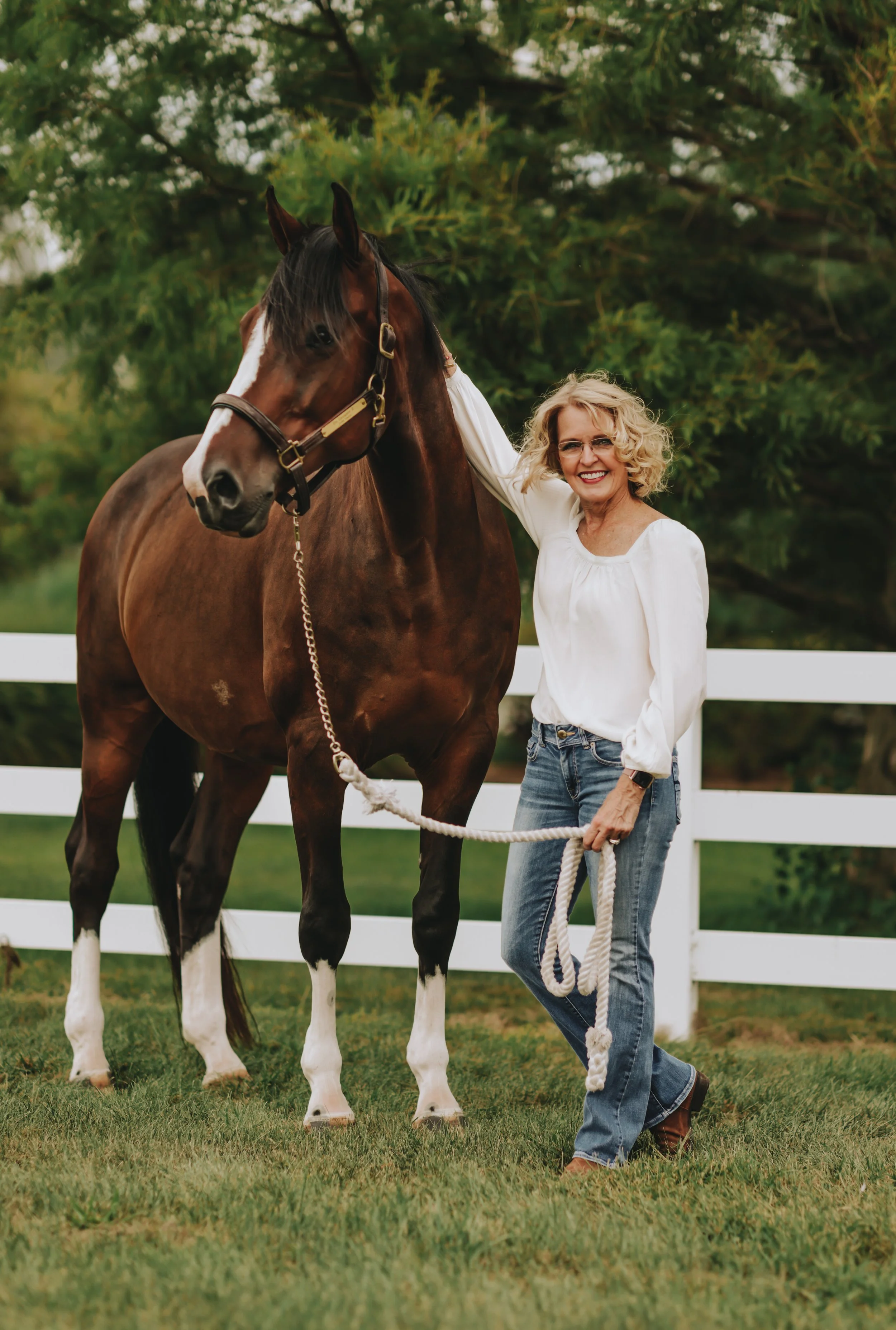 A woman with curly blonde hair, glasses, and a white blouse standing next to a brown and white horse in a grassy yard with a white fence and green trees in the background.