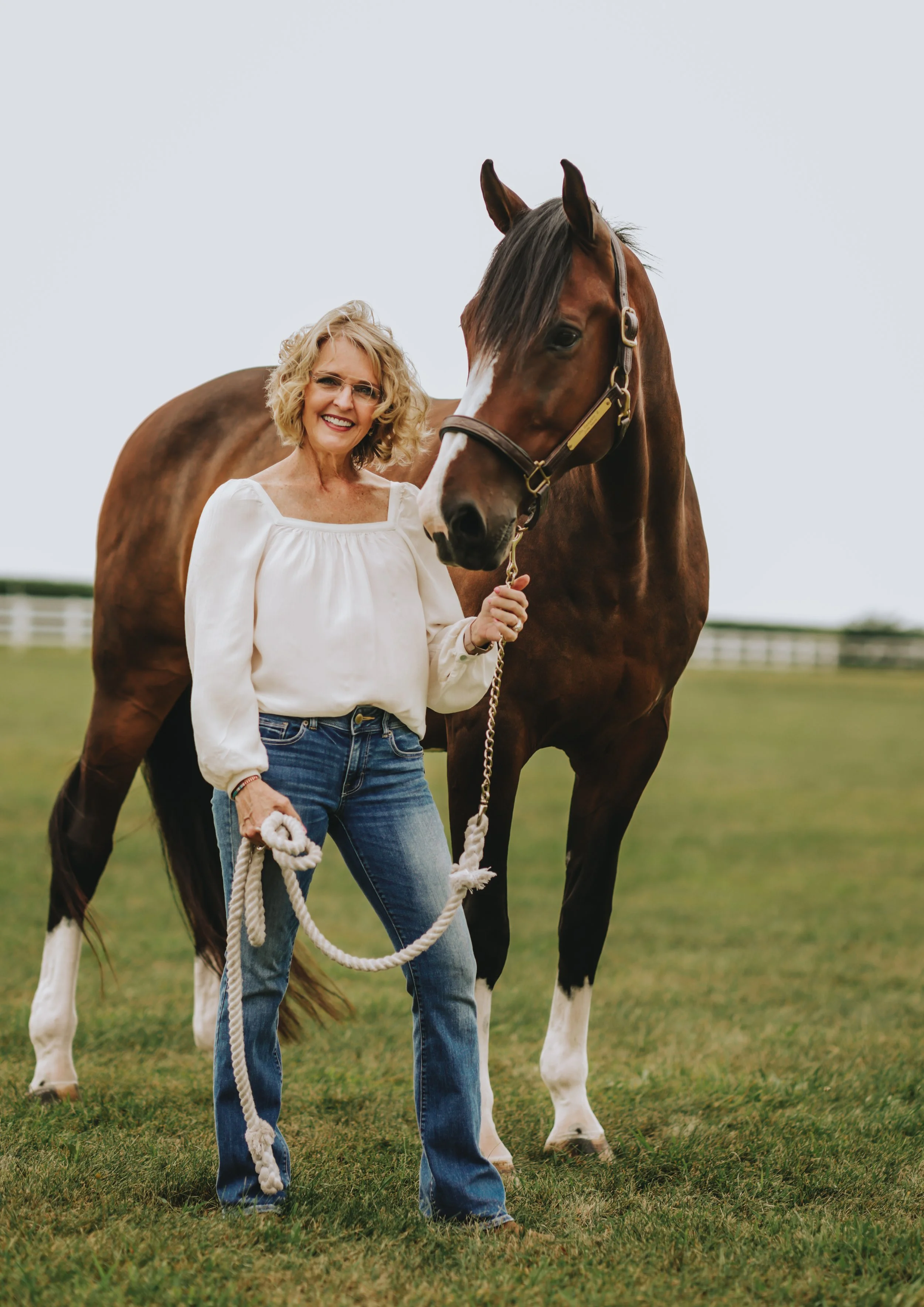 A smiling woman with curly blonde hair and glasses holding a lead rope while standing next to a brown and white horse on a grassy field with light gray sky in the background.