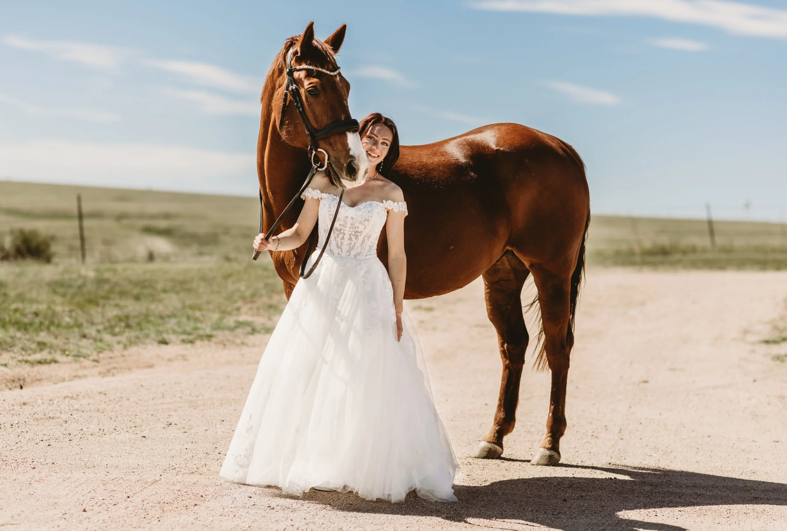 A woman in a wedding dress standing outdoors with a brown horse on a dirt path, under a blue sky with scattered clouds.