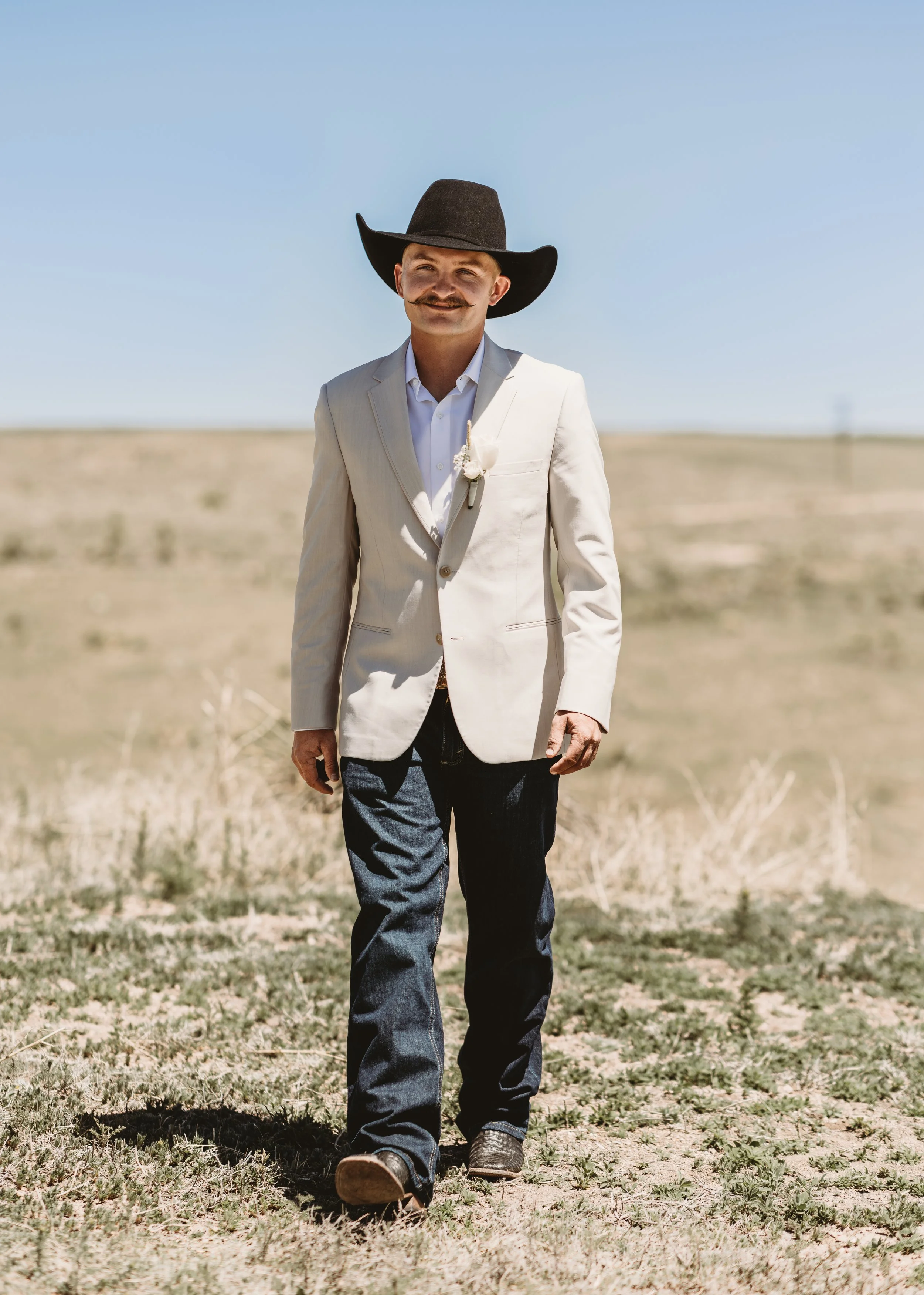 A man in a white blazer, jeans, and cowboy hat walking across a dry field on a sunny day