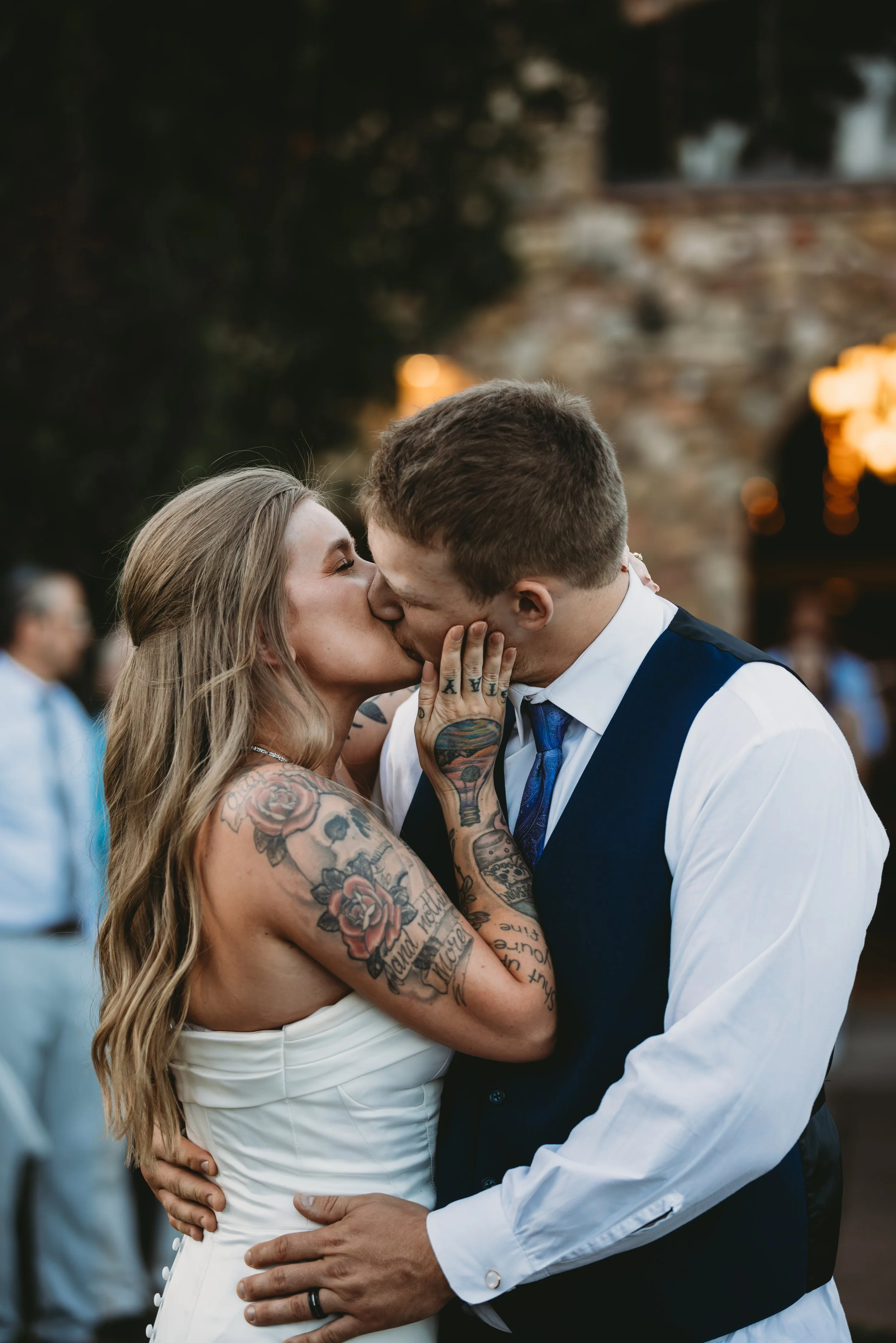 A bride and groom share a kiss during their wedding, with the bride's arms covered in colorful tattoos and the groom in a vest and tie, surrounded by blurred guests and cloudy sky.