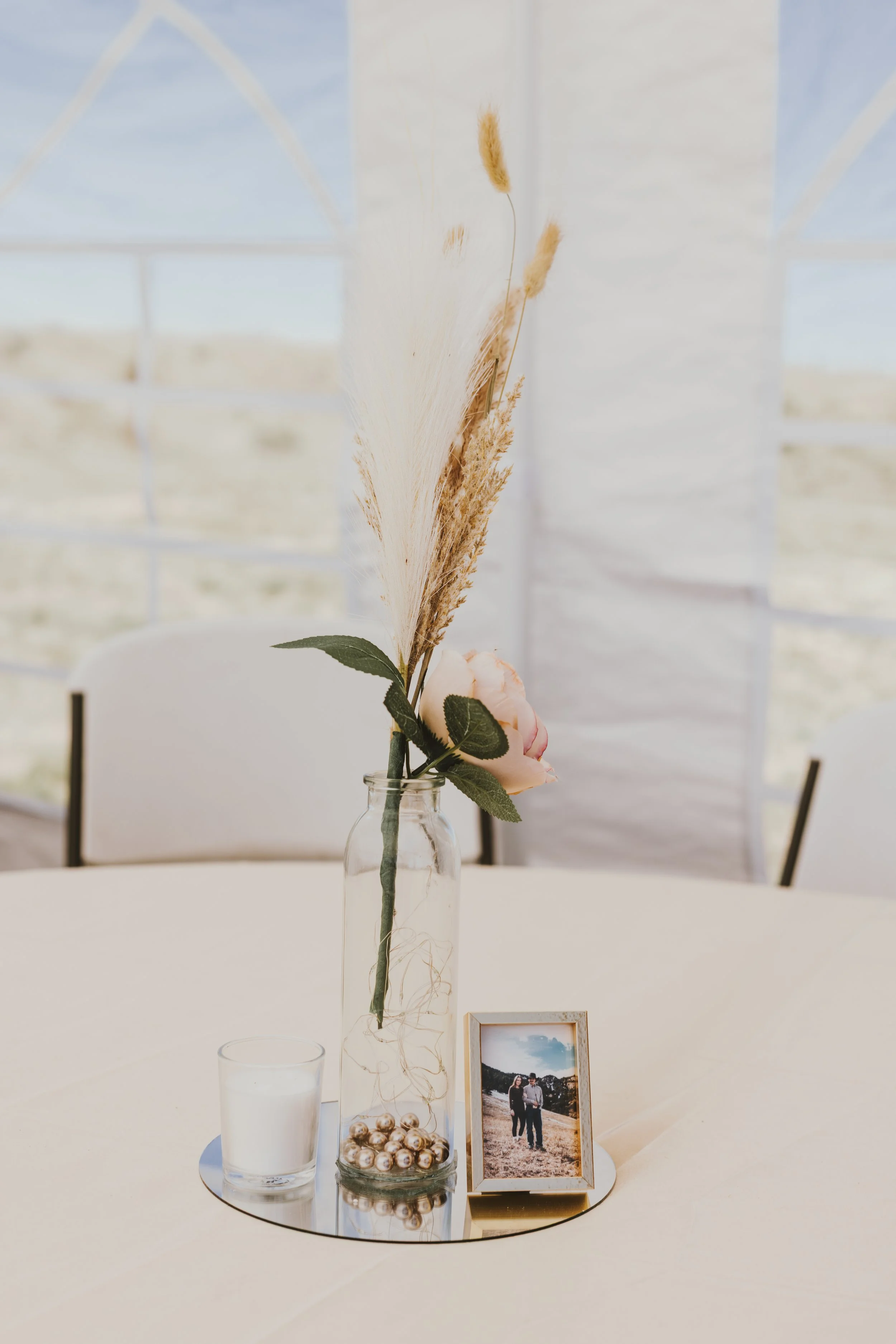 A table centerpiece with a clear glass vase holding pampas grass and roses, a white candle, and a framed photograph, all arranged on a circular mirror tray with decorative gold balls, set on a white table in a bright room.