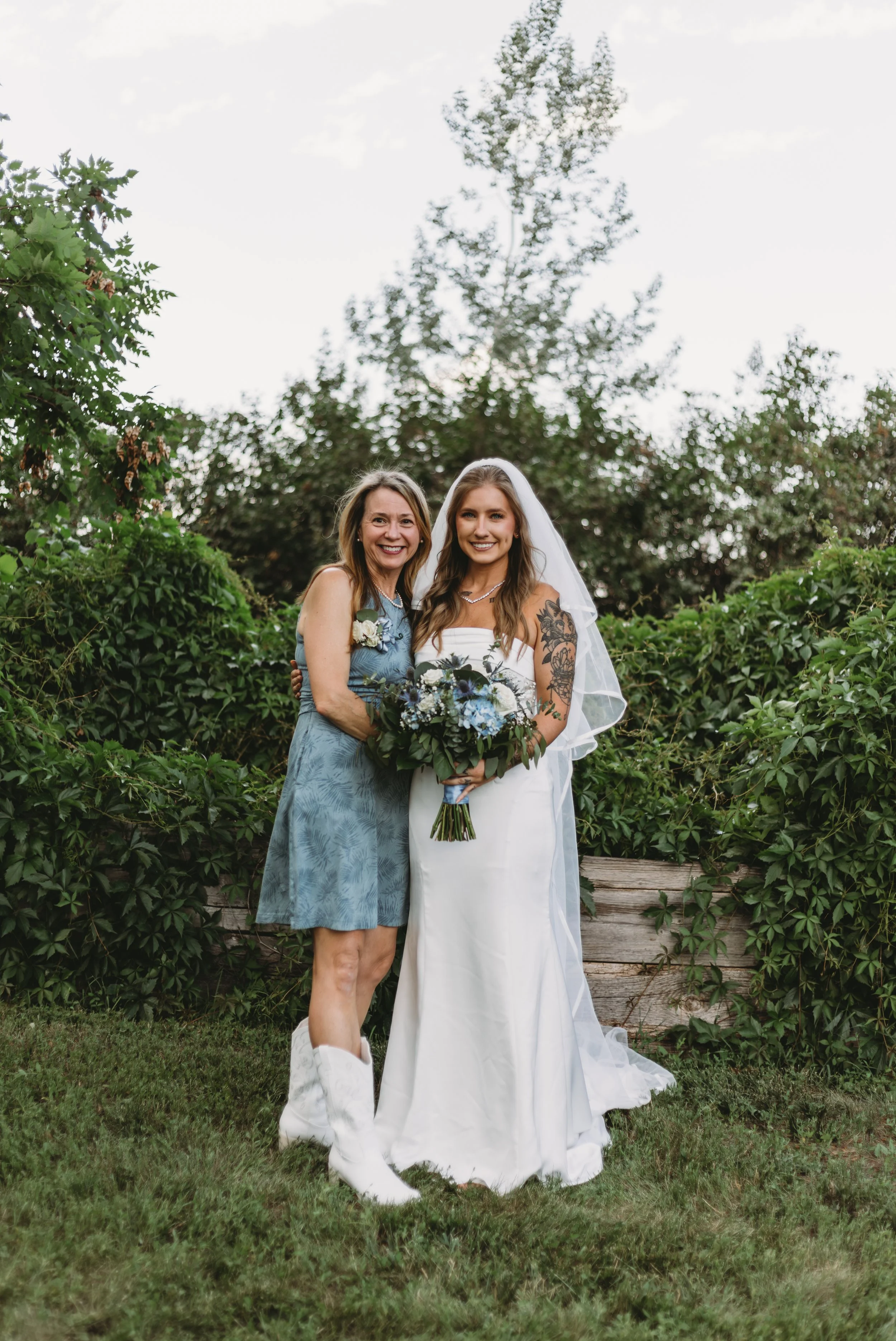 A bride and an older woman standing outdoors in front of green bushes and trees, smiling and posing together. The bride wears a white wedding dress and a veil, holding a bouquet of flowers. The older woman is dressed in a blue dress with white shoes 