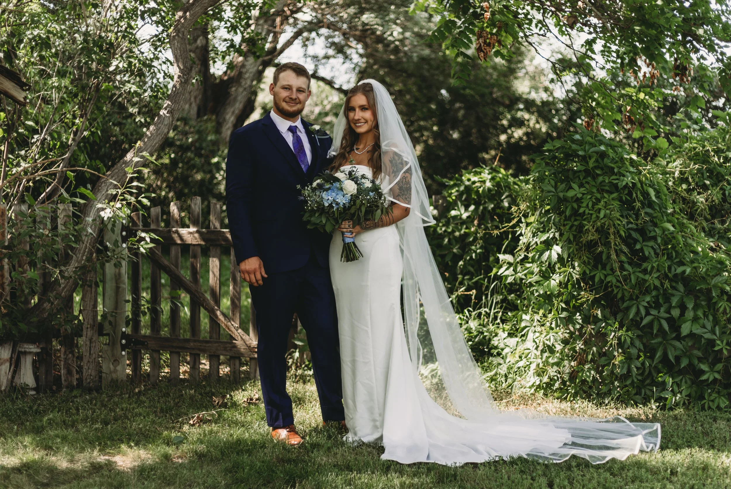 A newlywed couple stands outdoors in a lush, green setting. The groom is dressed in a navy suit with a white shirt and purple tie, while the bride wears a white wedding dress and veil, holding a bouquet of white and blue flowers. They are smiling and