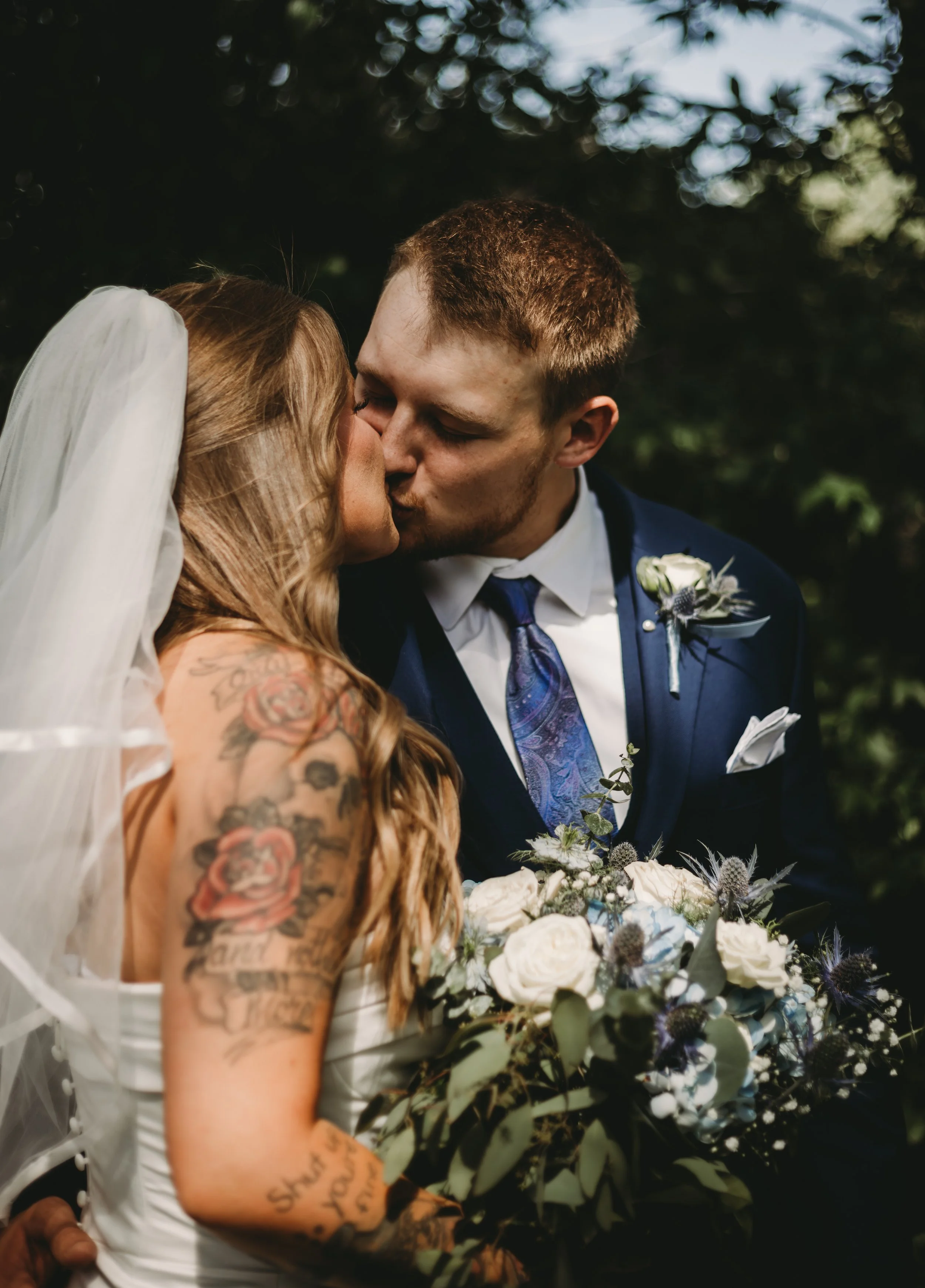 A bride and groom kissing outdoors during their wedding ceremony, with the bride's tattoos visible on her arm and bouquet of white flowers in front of them.