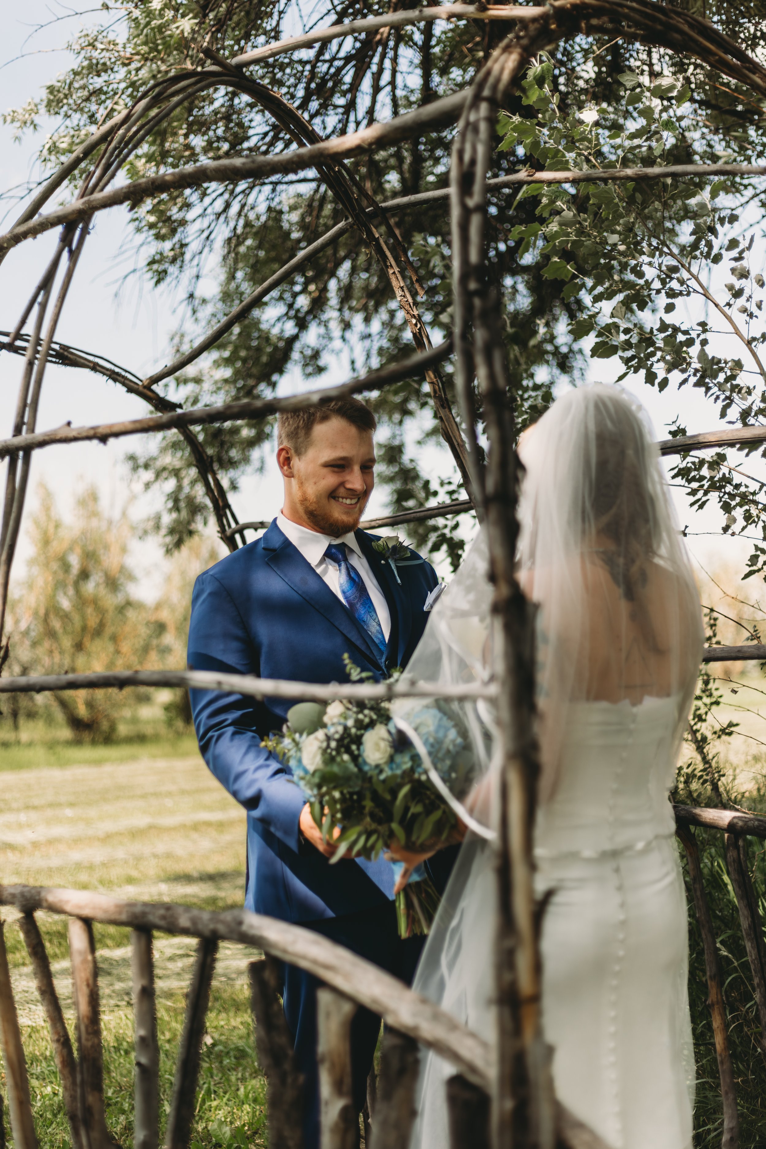 A bride and groom exchanging vows outdoors during a wedding ceremony, viewed through a rustic wooden archway with greenery and trees in the background.