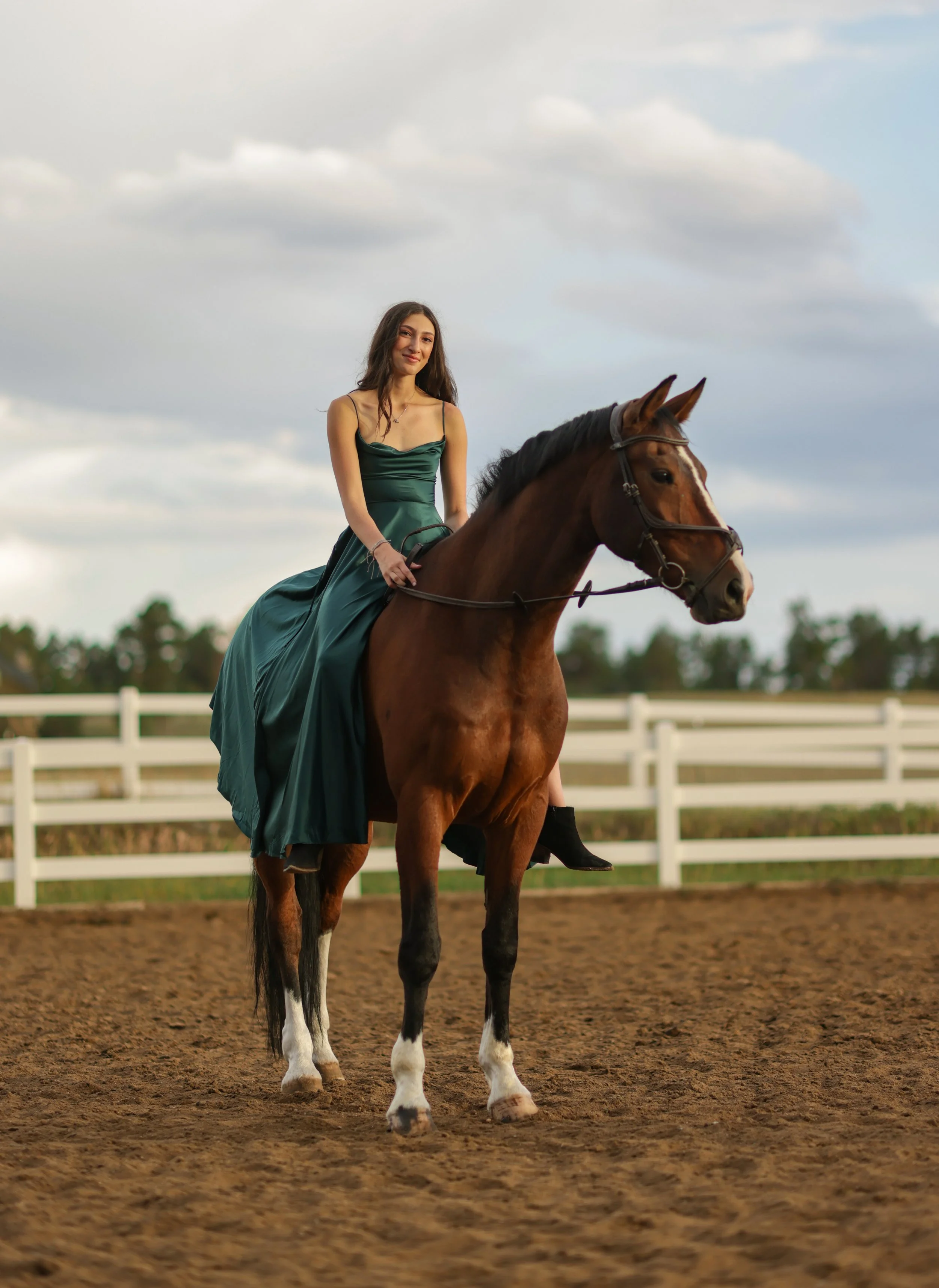 A woman in a green dress riding a brown horse with white markings on its legs, in an enclosed outdoor riding arena with a white fence, under a cloudy sky.