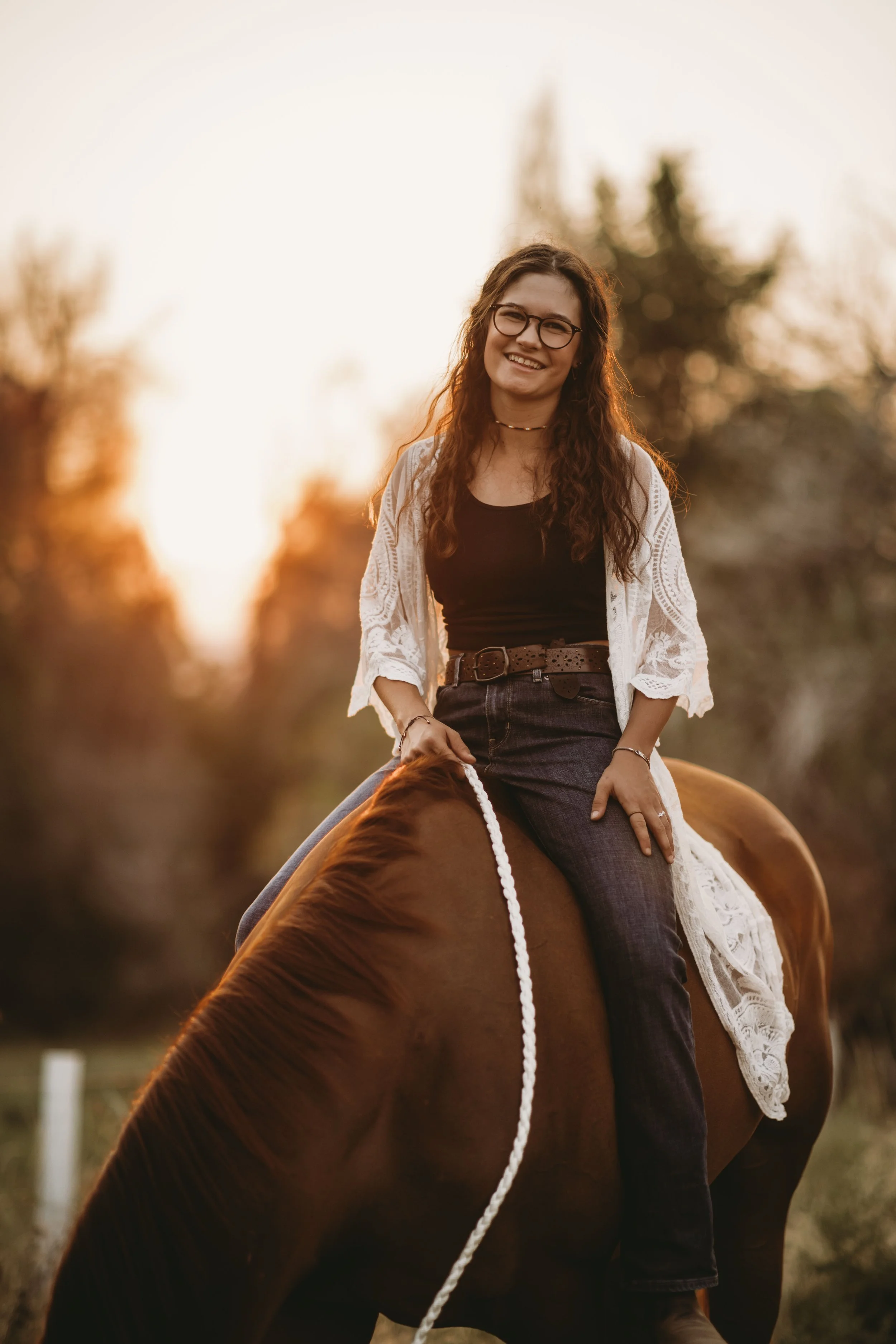 Young woman with glasses riding a brown horse during sunset, smiling, outdoors with trees in the background.