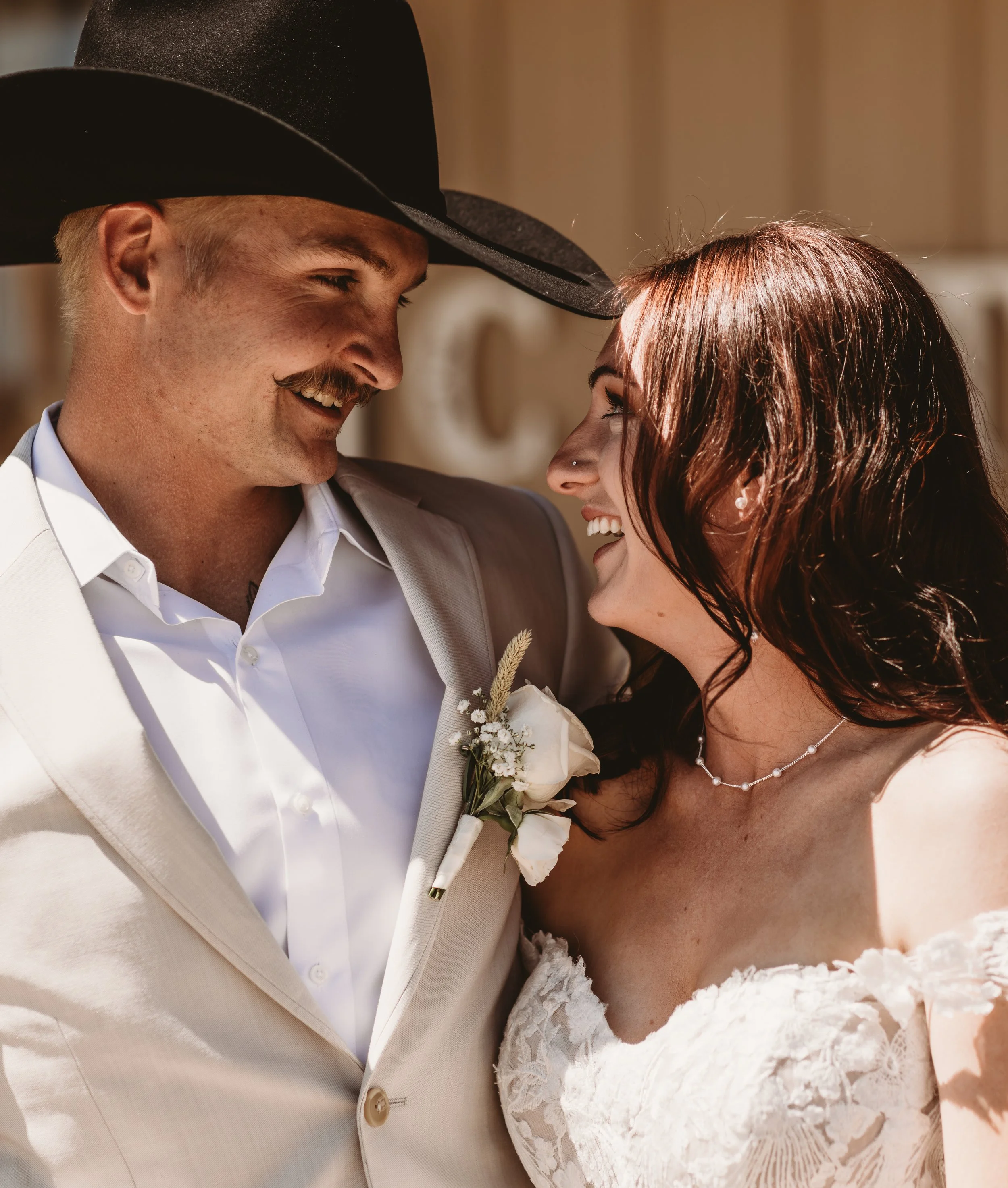 Close-up of a bride and groom smiling and gazing at each other during their wedding, with the groom wearing a light beige suit and black hat, and the bride in a white lace dress with a pearl necklace.