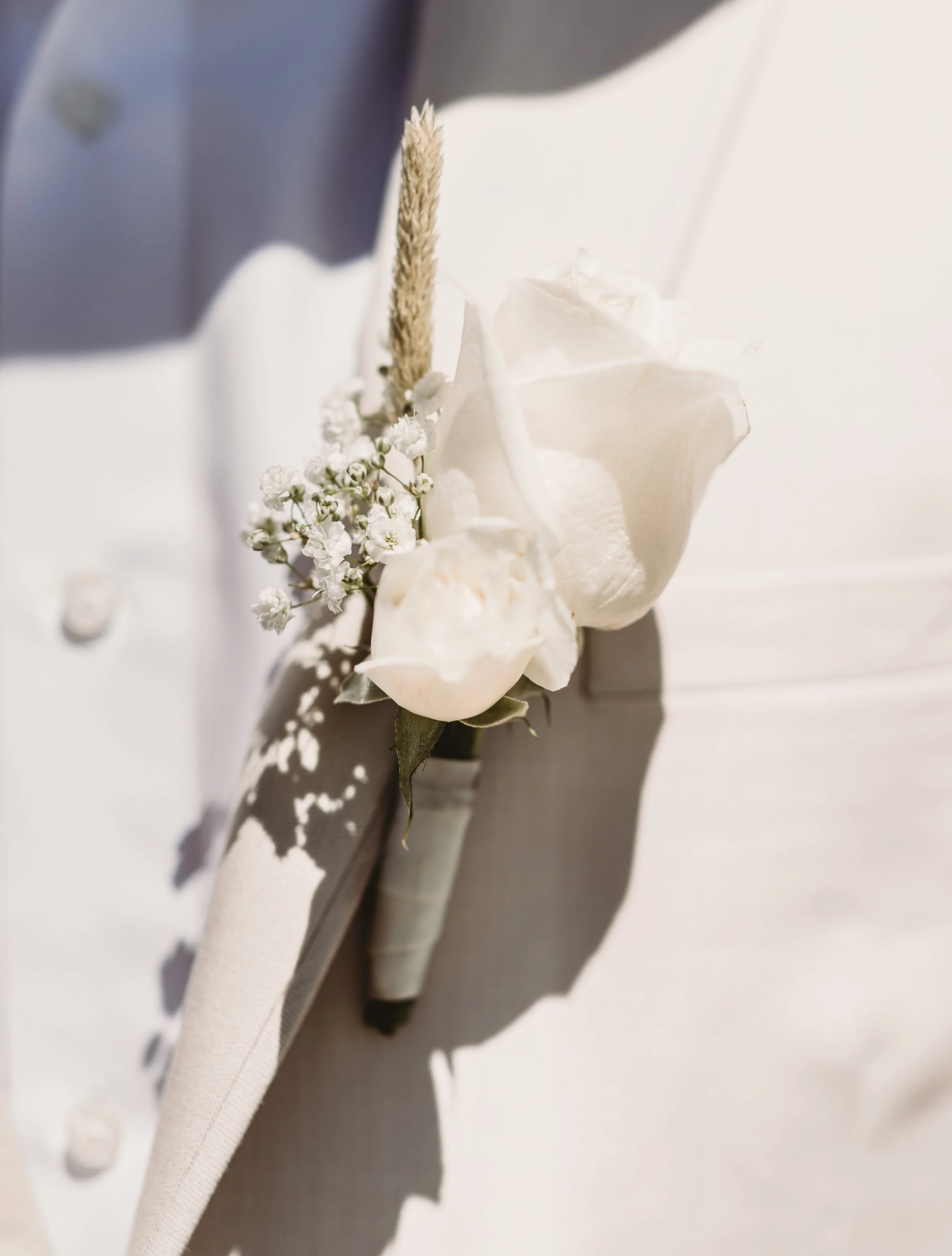 Close-up of a white flower corsage with baby's breath, a dried grass element, and a white ribbon, attached to a piece of white or cream-colored fabric.