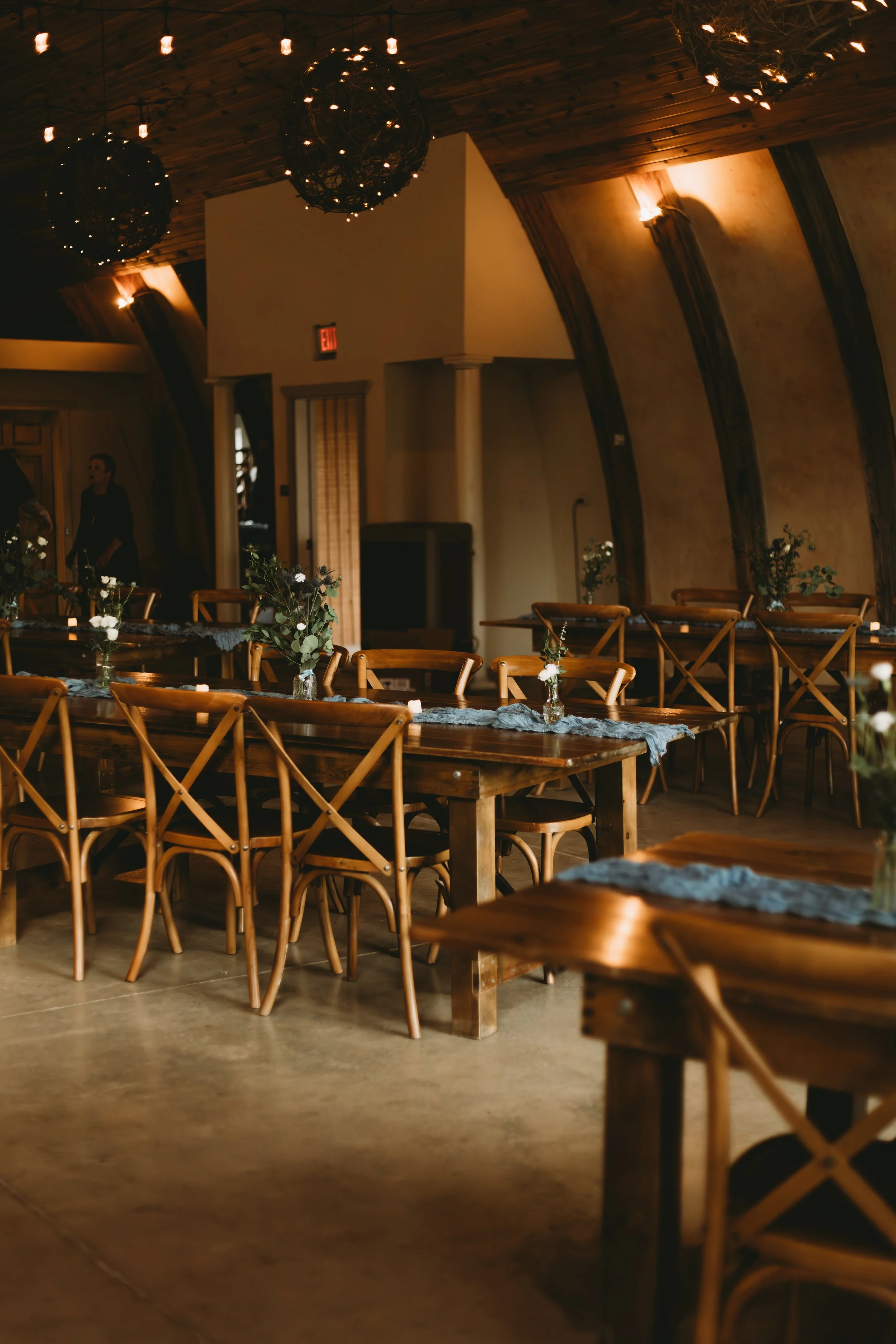 Interior view of a decorated event or dining space with wooden tables, chairs, and small flower arrangements; illuminated by hanging and wall lights, with a wooden ceiling and curved wall design.