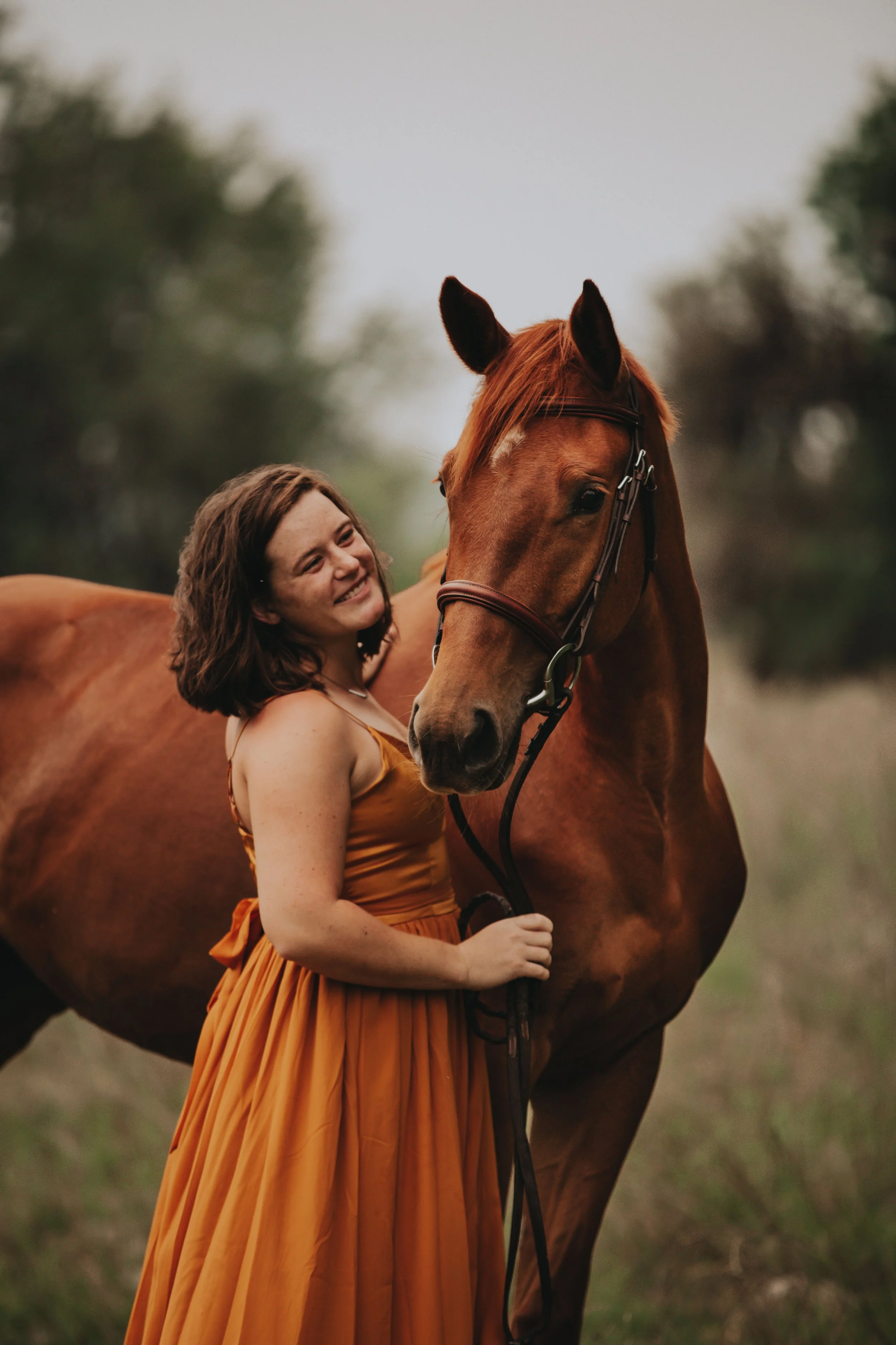 A woman in an orange dress hugging a brown horse outdoors with trees in the background.