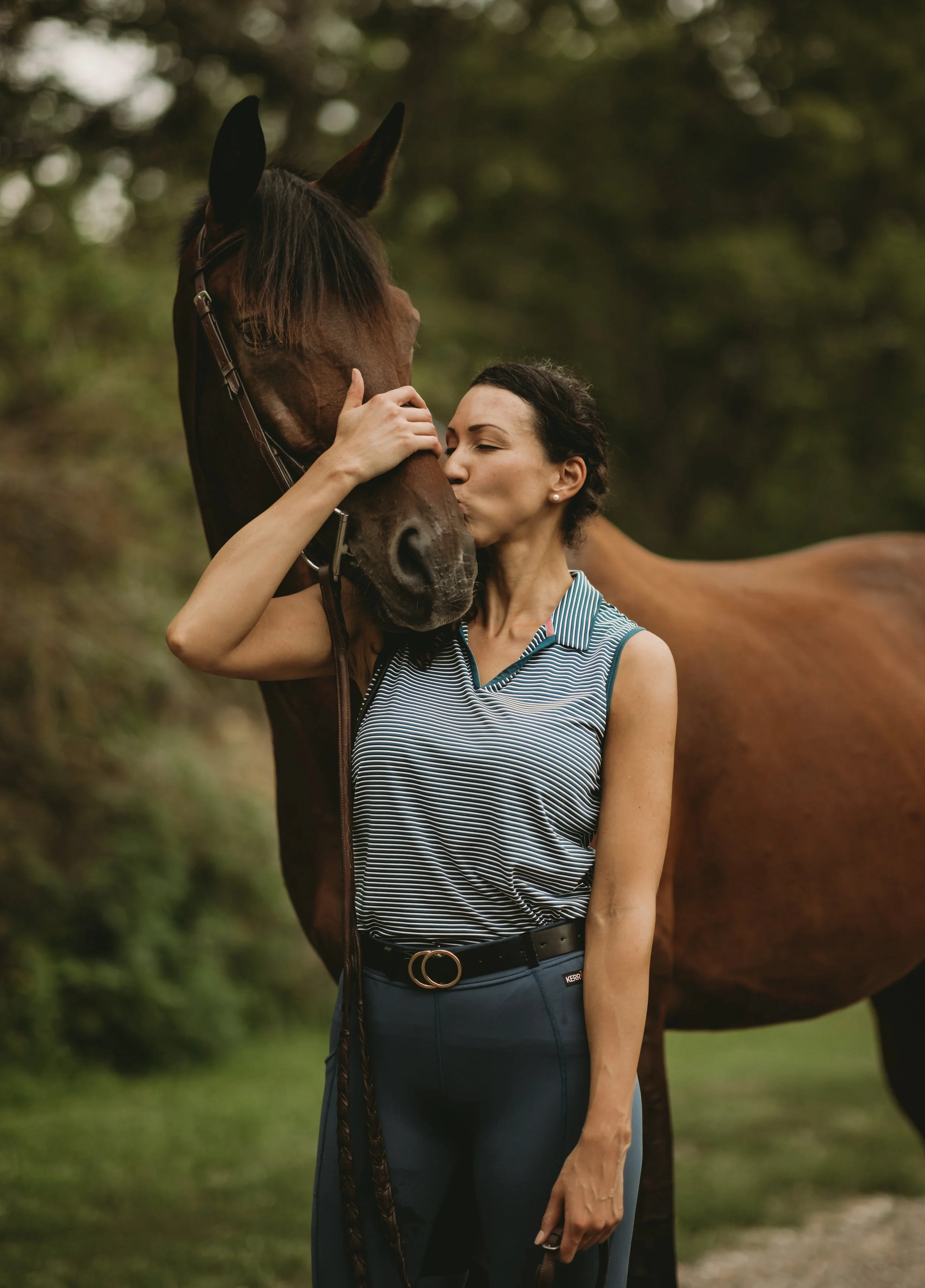 A woman kissing a horse on its face outdoors, surrounded by trees.