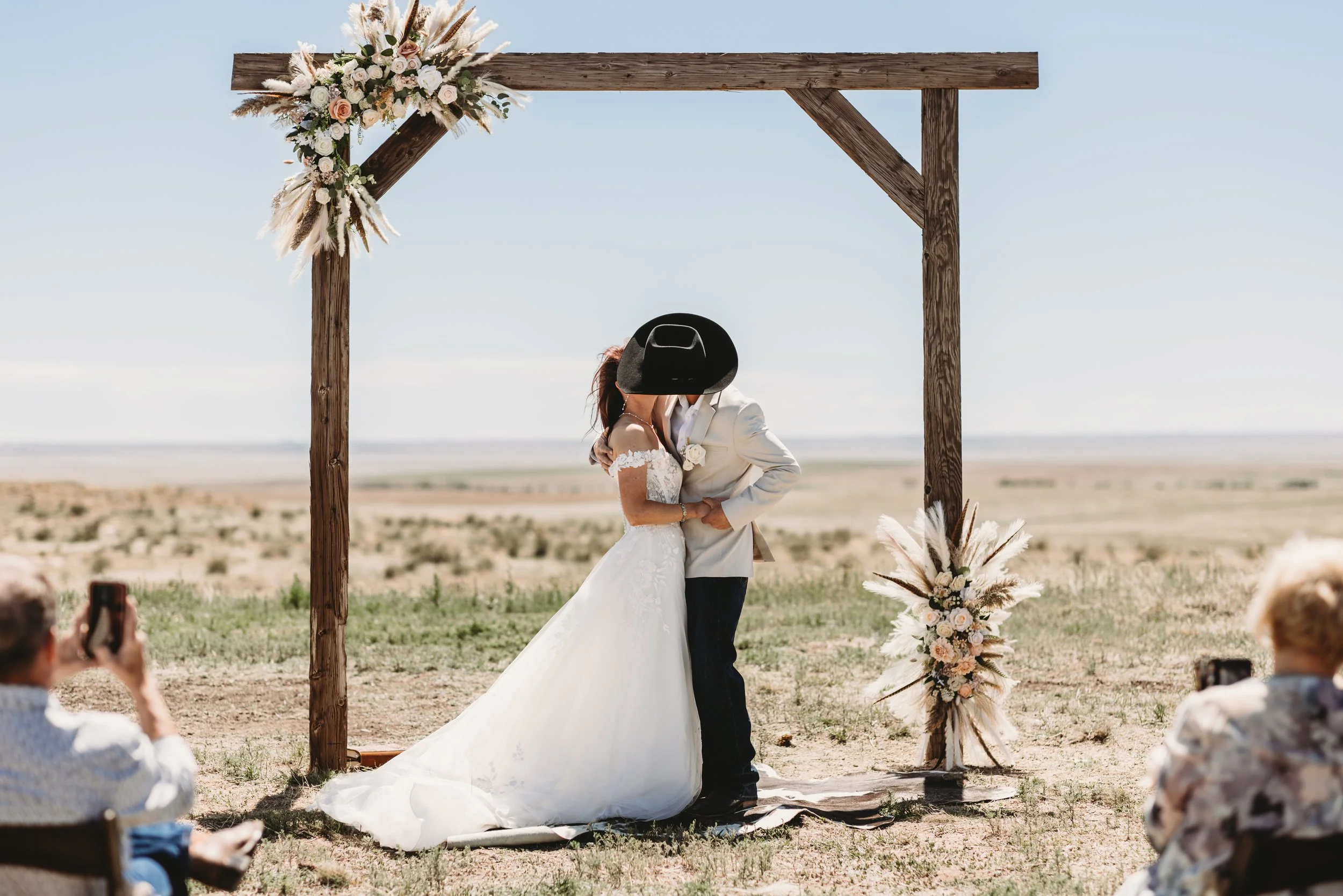 A bride and groom kissing during their outdoor wedding ceremony under a wooden arch decorated with flowers, set in a desert landscape with guests seated and taking photos.