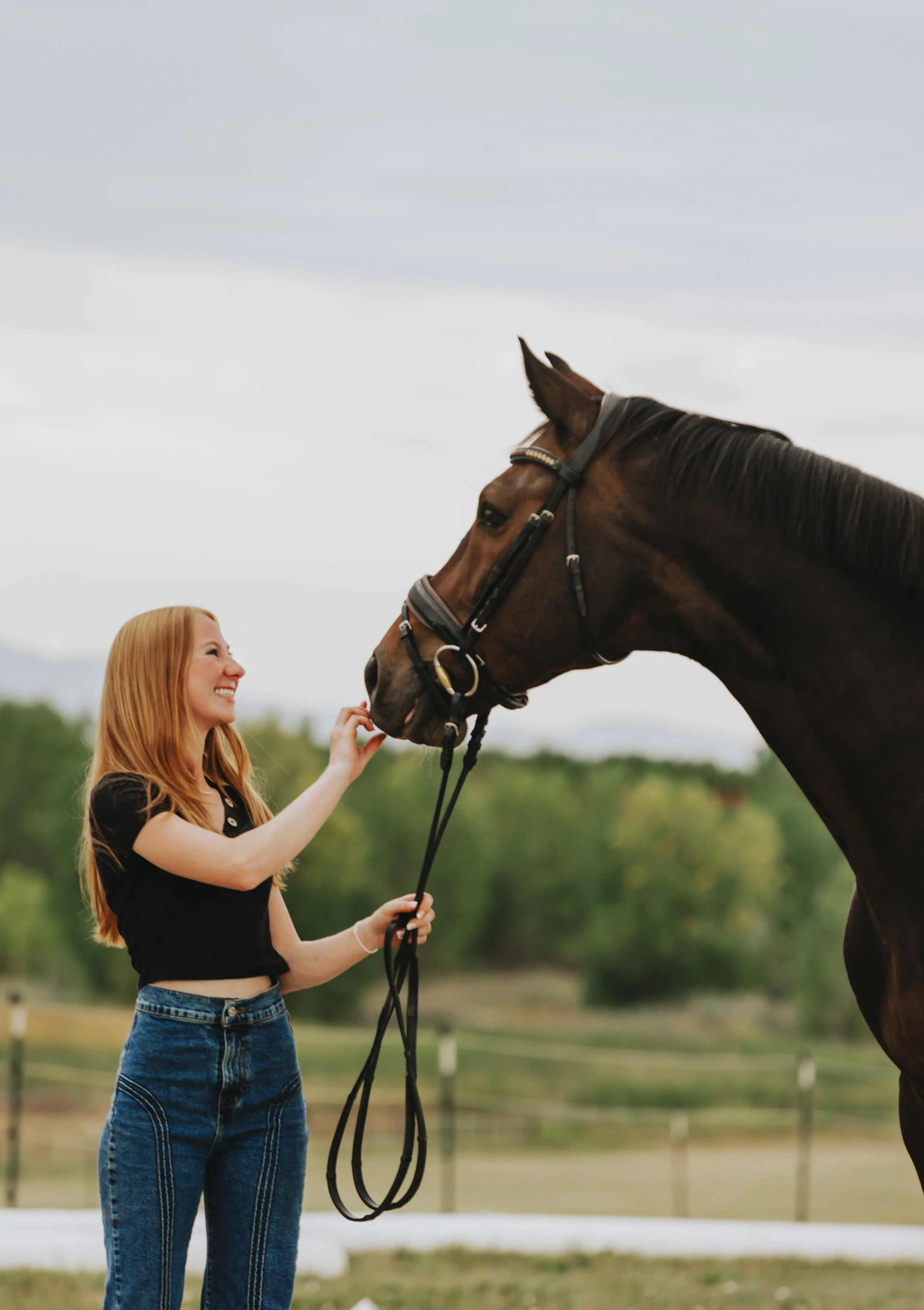 A young woman with red hair smiling and holding the reins of a brown horse outdoors with a cloudy sky and greenery in the background.