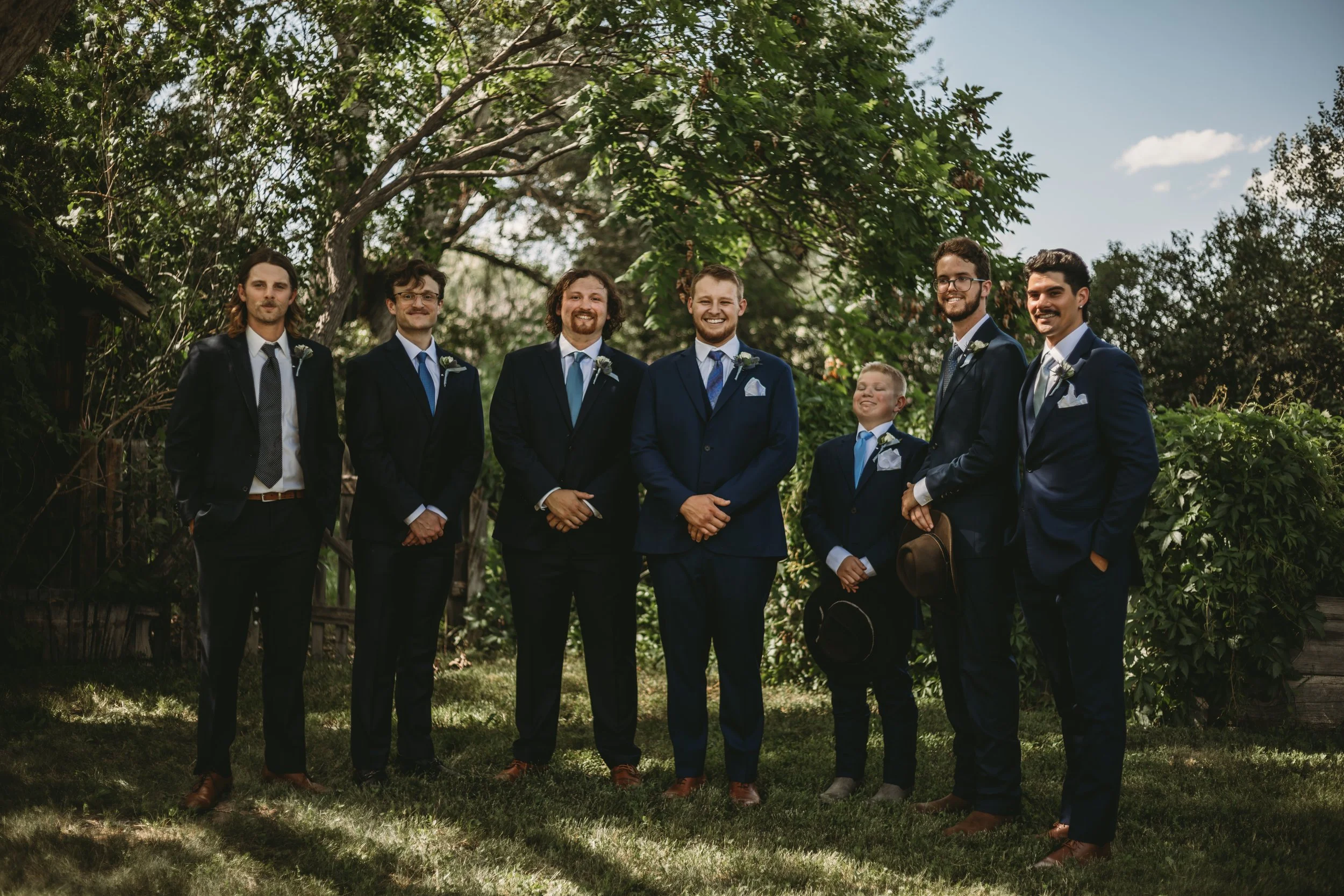 A group of seven men dressed in formal suits, standing outdoors under trees with green foliage. They are smiling and appear to be at a wedding or formal event, with some wearing boutonnieres and holding hats.