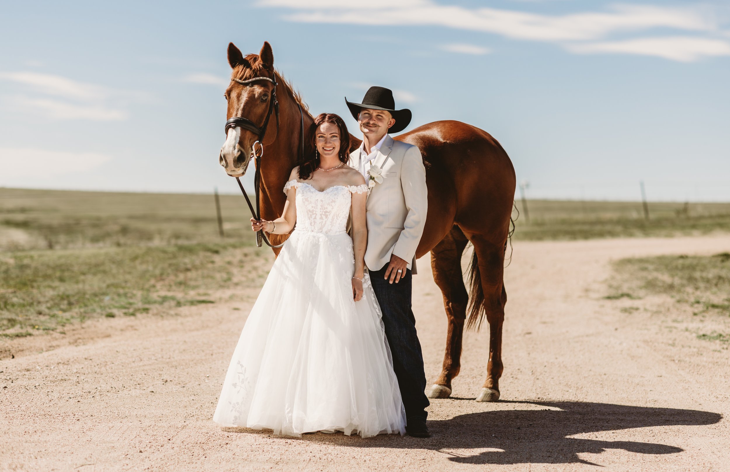 A bride in a white wedding dress standing next to a man in a white suit and black cowboy hat, holding a rearing brown horse in a rural outdoor setting under a partly cloudy sky.