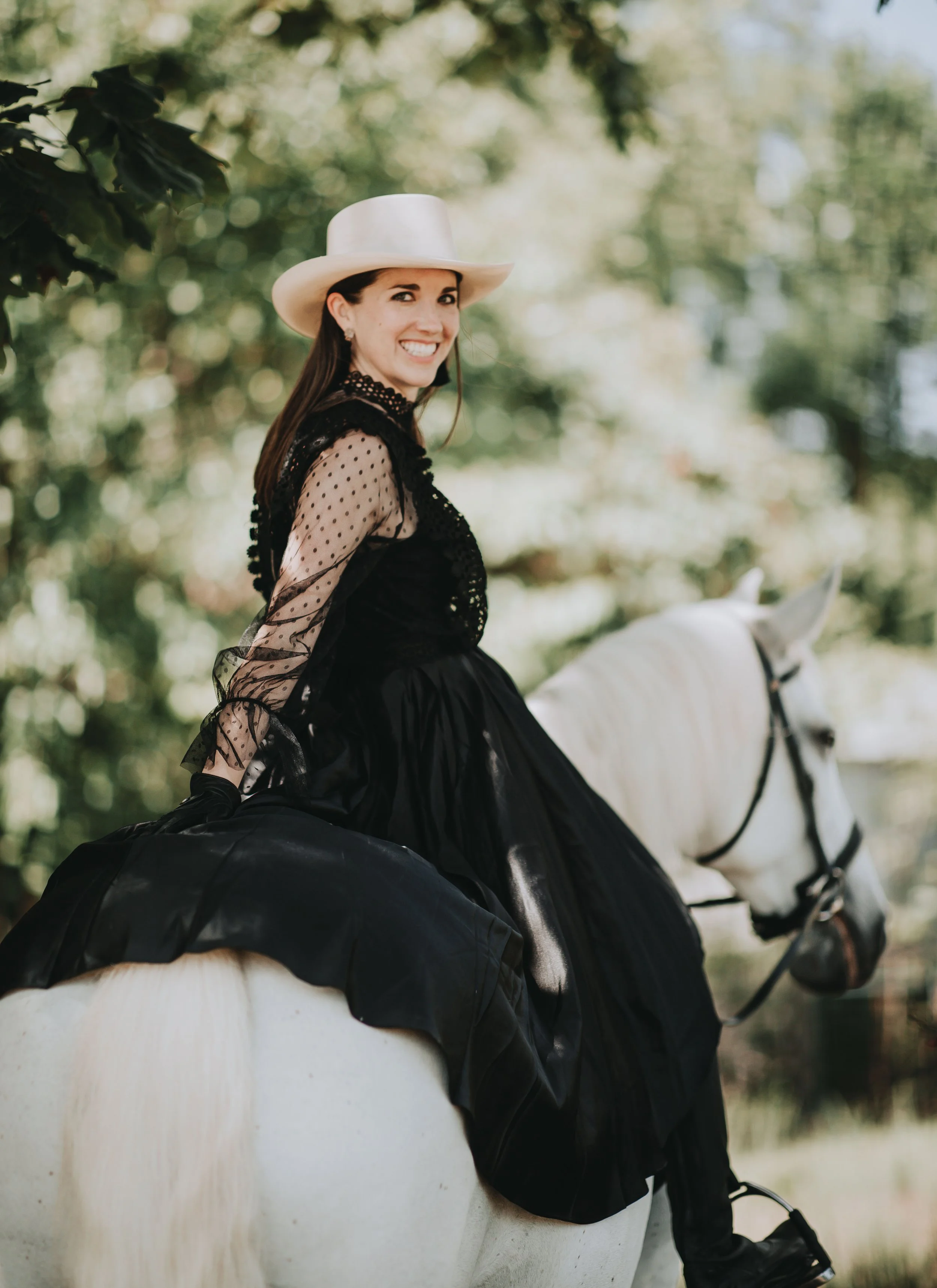 Woman in a black dress and a white hat riding a white horse outdoors surrounded by green trees.
