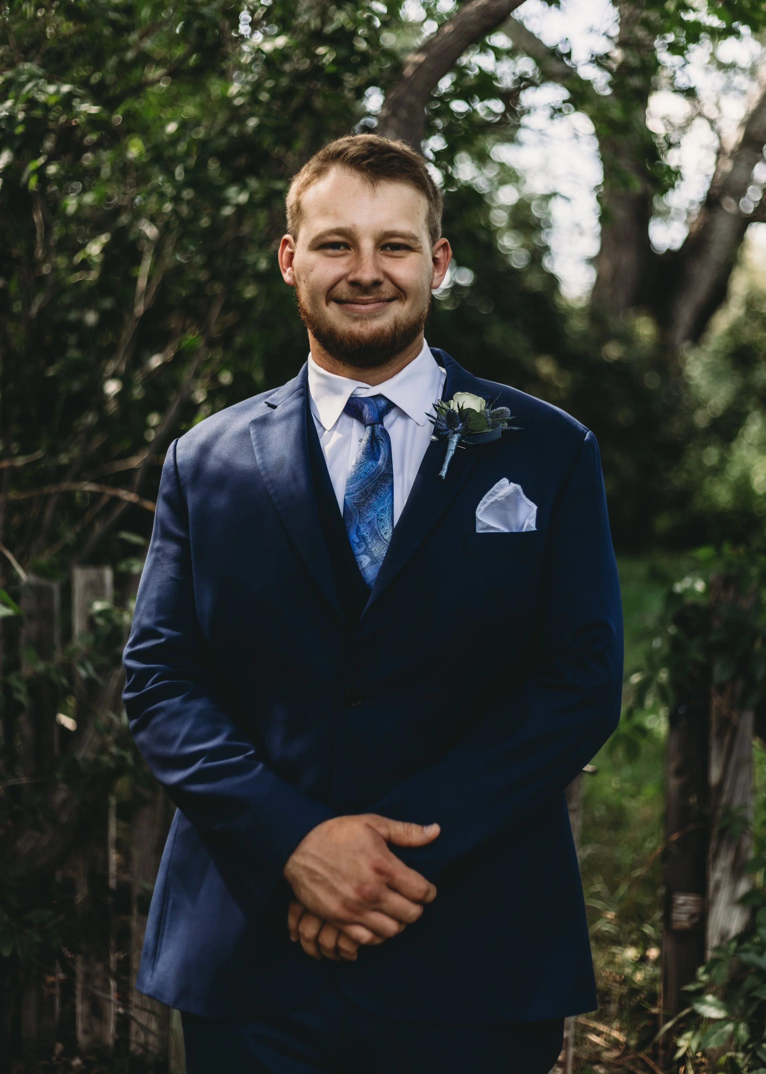 A man with light brown hair and a beard wearing a dark blue suit, white shirt, and a blue patterned tie, standing outdoors in front of green trees and foliage, smiling at the camera.