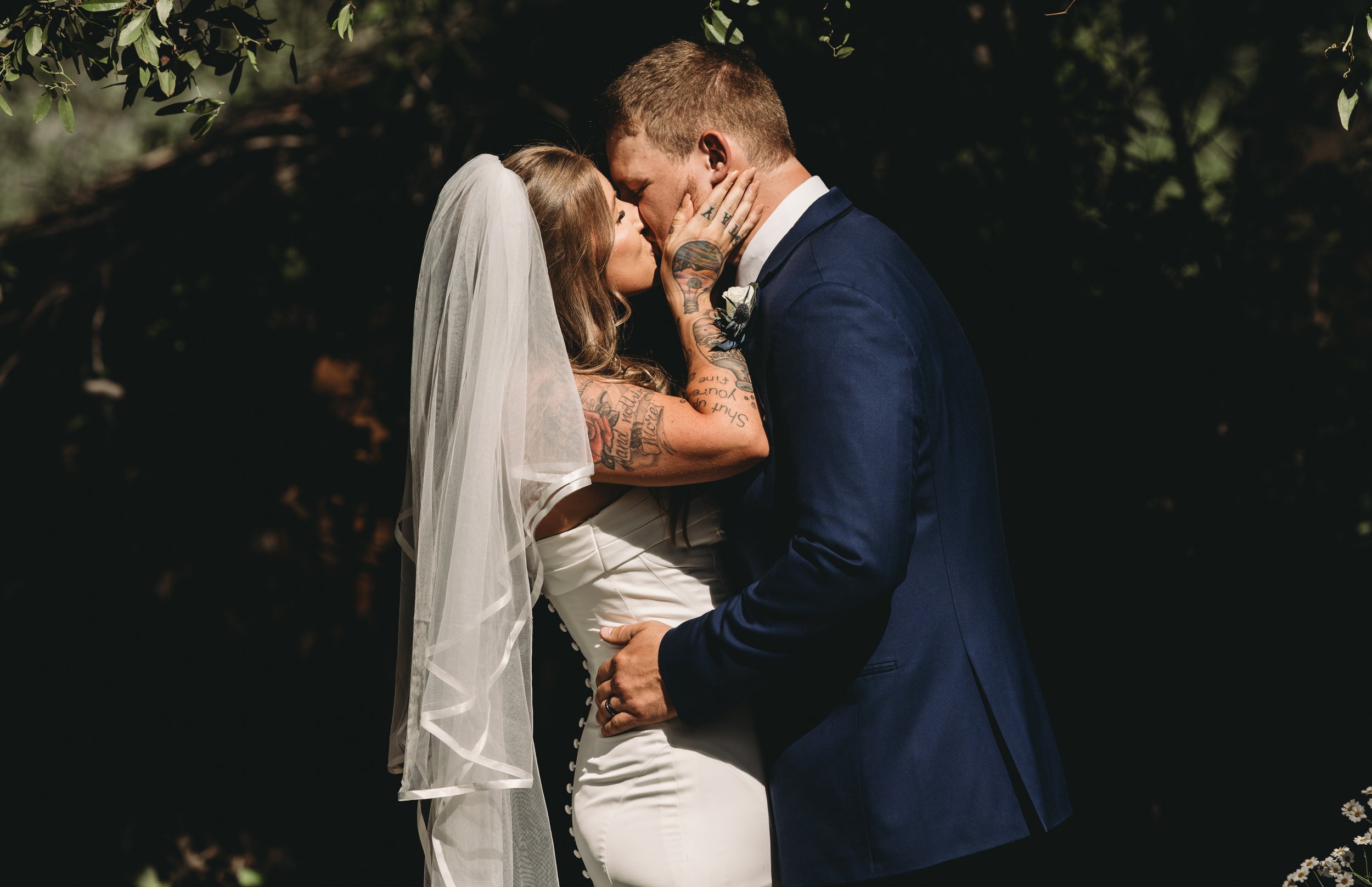A bride and groom share a kiss during their outdoor wedding ceremony, with greenery in the background. The bride has long blonde hair, a white veil, and tattoos on her arms, while the groom wears a dark blue suit and has short hair.
