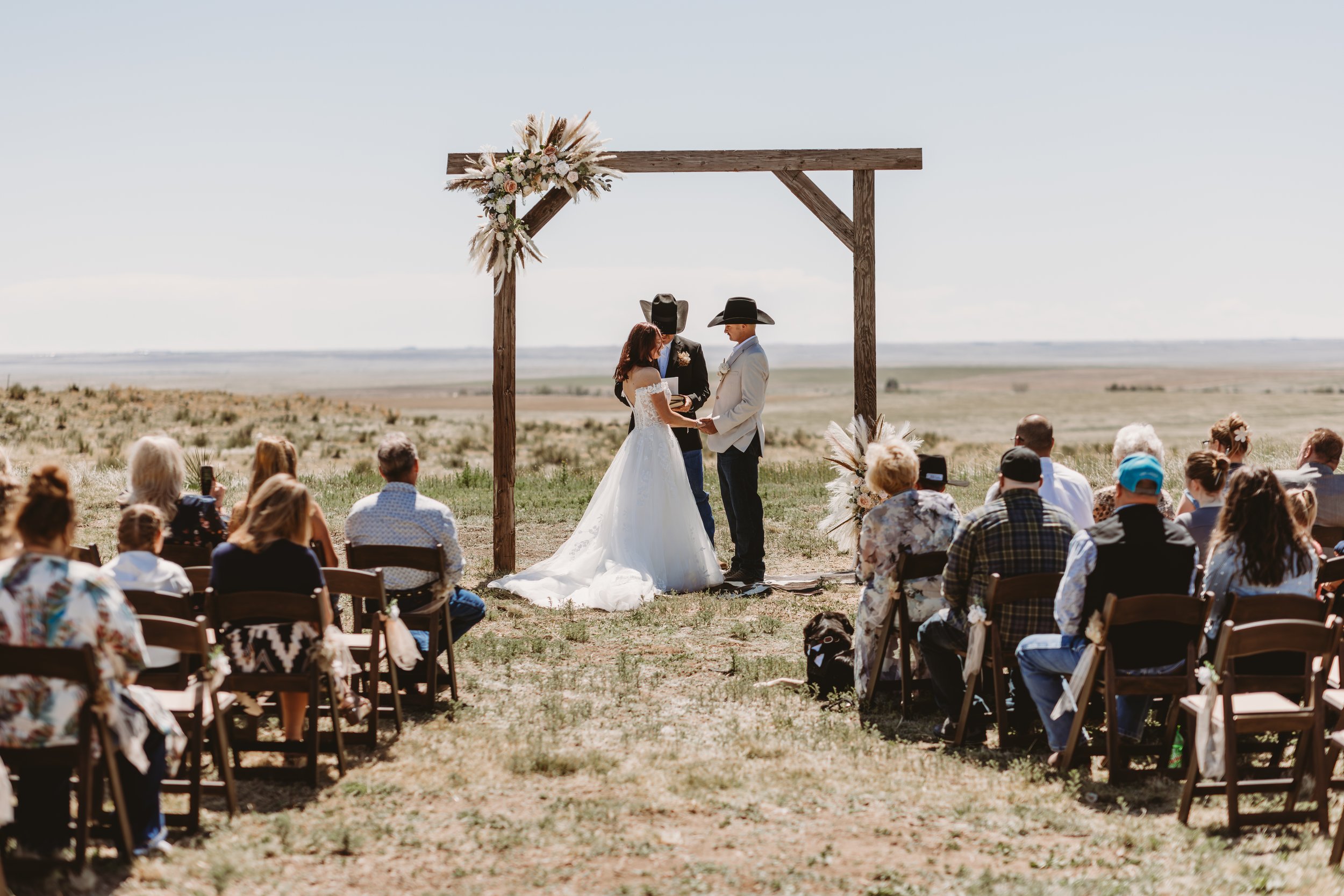 A couple getting married outdoors under a wooden arch decorated with flowers, with guests seated on both sides as a officiant conducts the ceremony.
